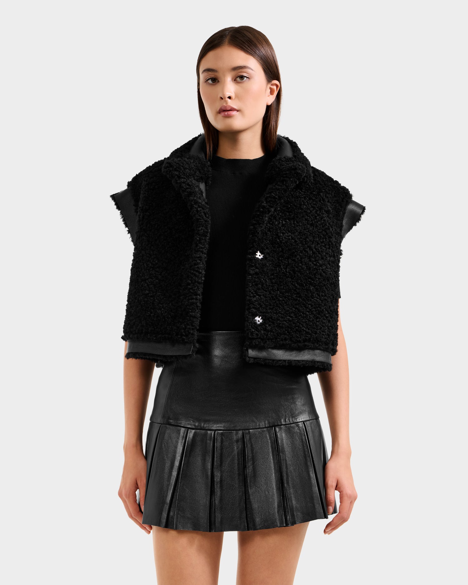 A woman with straight brown hair and a neutral expression wears the Rudsak PALOMA - 8125675 Black textured sleeveless vest over a black top and pleated leather skirt, standing against a plain light background.