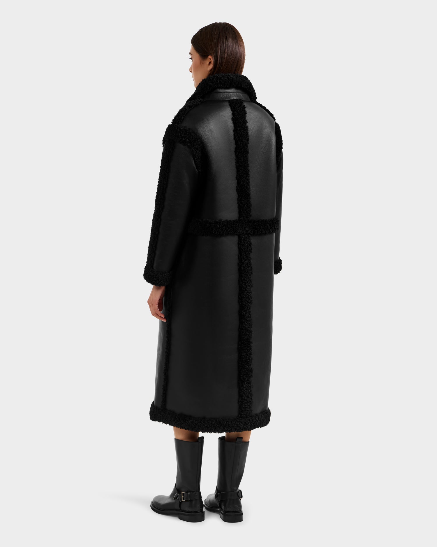 A person faces away, wearing the Rudsak PALOMA - 8125675 Black long faux shearling coat with textured trim and black boots. The reversible women’s coat features a straight, oversized fit and is paired with straight brown hair past the shoulders.