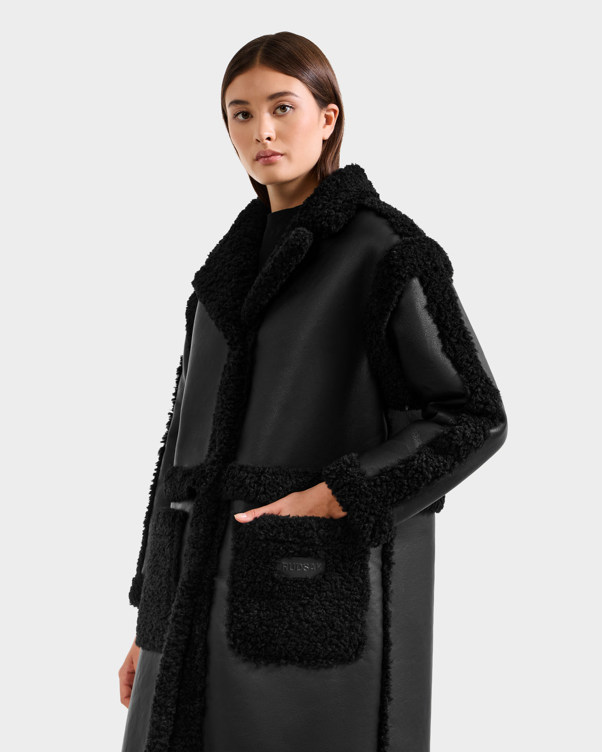 A woman models the Rudsak PALOMA - 8125675 Black faux shearling coat with textured trim and large front pockets, posing against a plain light gray background.
