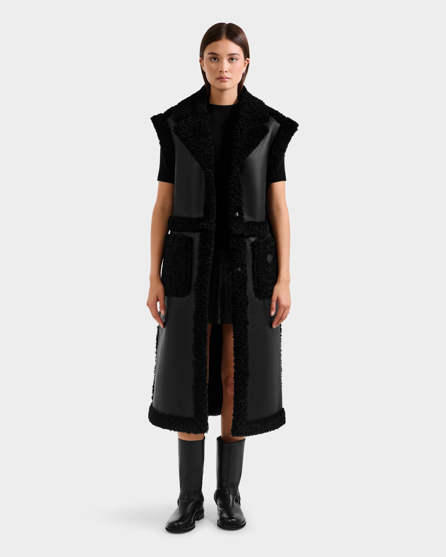 A woman faces forward against a plain background, wearing the Rudsak PALOMA - 8125675 Black long sleeveless faux shearling coat with textured trim, paired with a black top, skirt, and knee-high boots.