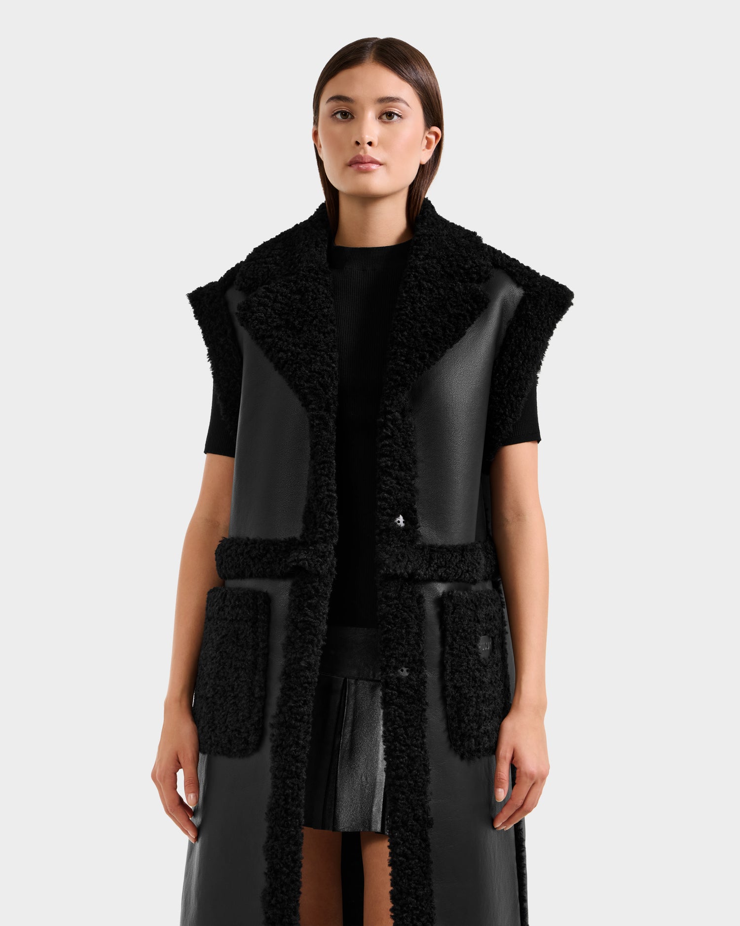 A woman faces forward, wearing the Rudsak PALOMA - 8125675 Black sleeveless faux shearling vest with textured trim over a black top and skirt, set against a plain light background.