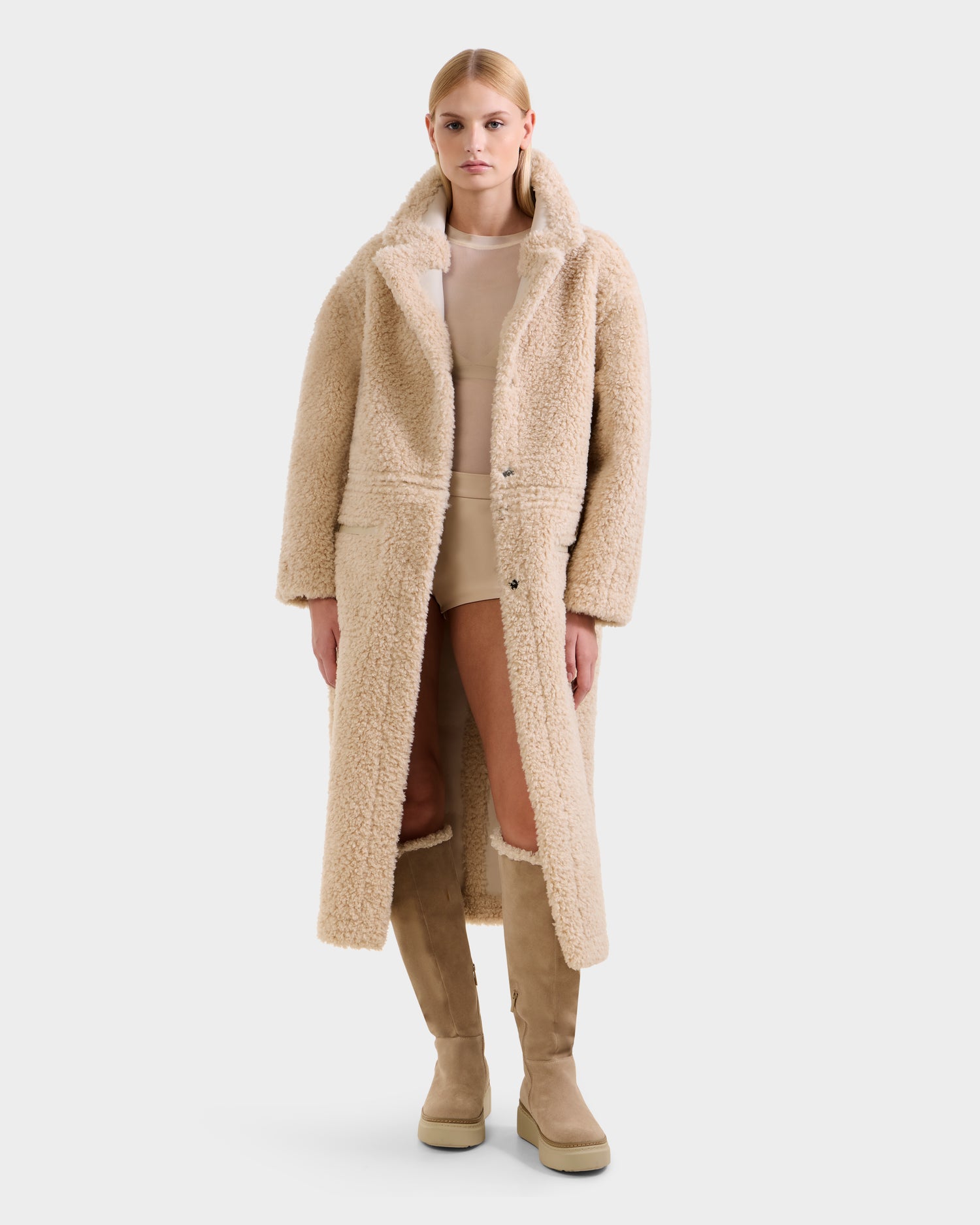 A woman stands facing forward against a white background, wearing the Rudsak PALOMA - 8125675 Vanilla faux shearling coat over a light beige outfit and matching knee-high boots.