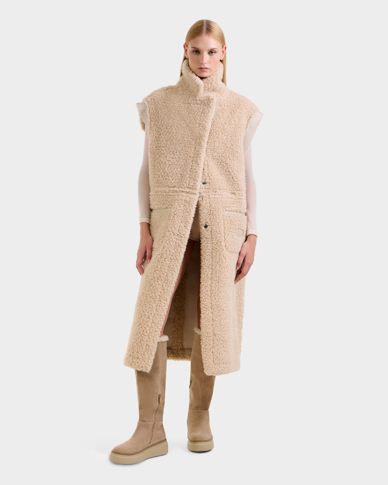 A person wears the Rudsak PALOMA - 8125675 Vanilla, a long, sleeveless faux shearling vest over a light top with matching beige knee-high boots, showcasing modular outerwear's versatile styling against a plain white background.
