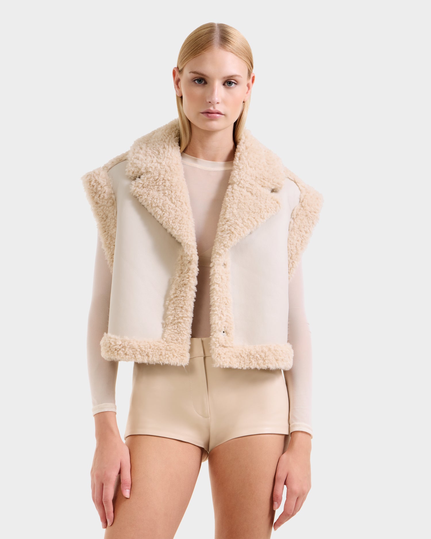 A woman models the Rudsak PALOMA - 8125675 Vanilla faux shearling vest over a sheer long-sleeve top and high-waisted beige shorts, standing against a plain white background and looking directly at the camera.