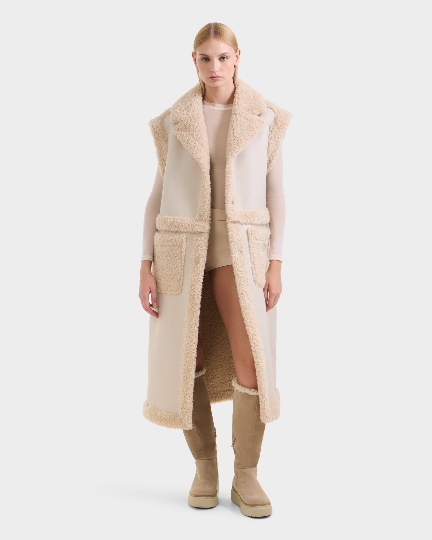 A woman faces forward against a white background wearing the Rudsak PALOMA - 8125675 Vanilla long, sleeveless, cream-colored faux shearling vest over a light beige top and shorts, paired with tan knee-high boots.