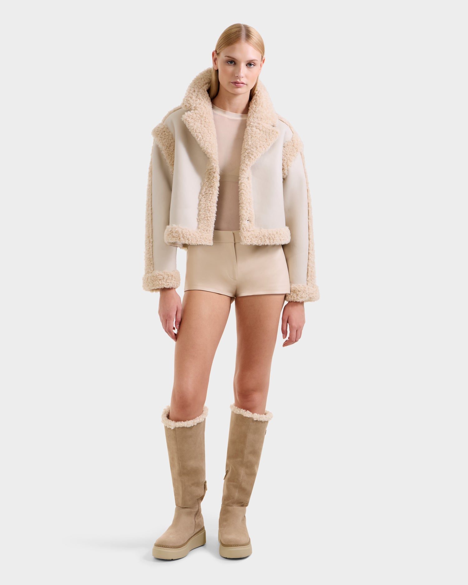 A woman with straight blonde hair and a neutral expression wears the Rudsak PALOMA - 8125675 Vanilla faux shearling jacket against a plain background, styled with a sheer beige top, beige shorts, and tall beige shearling boots.