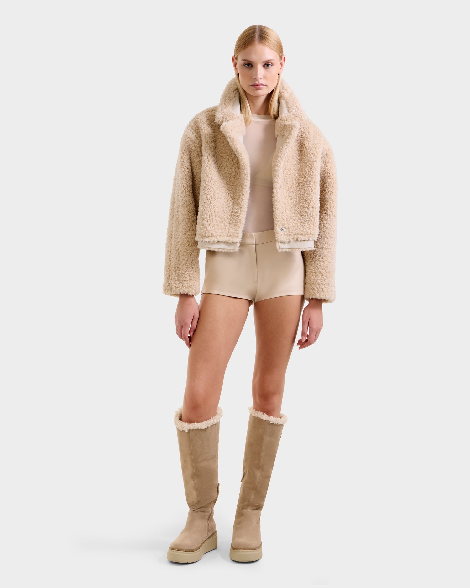 A woman faces forward in a Rudsak PALOMA - 8125675 Vanilla faux shearling sherpa jacket, beige top, shorts, and tall beige shearling boots against a plain white background.