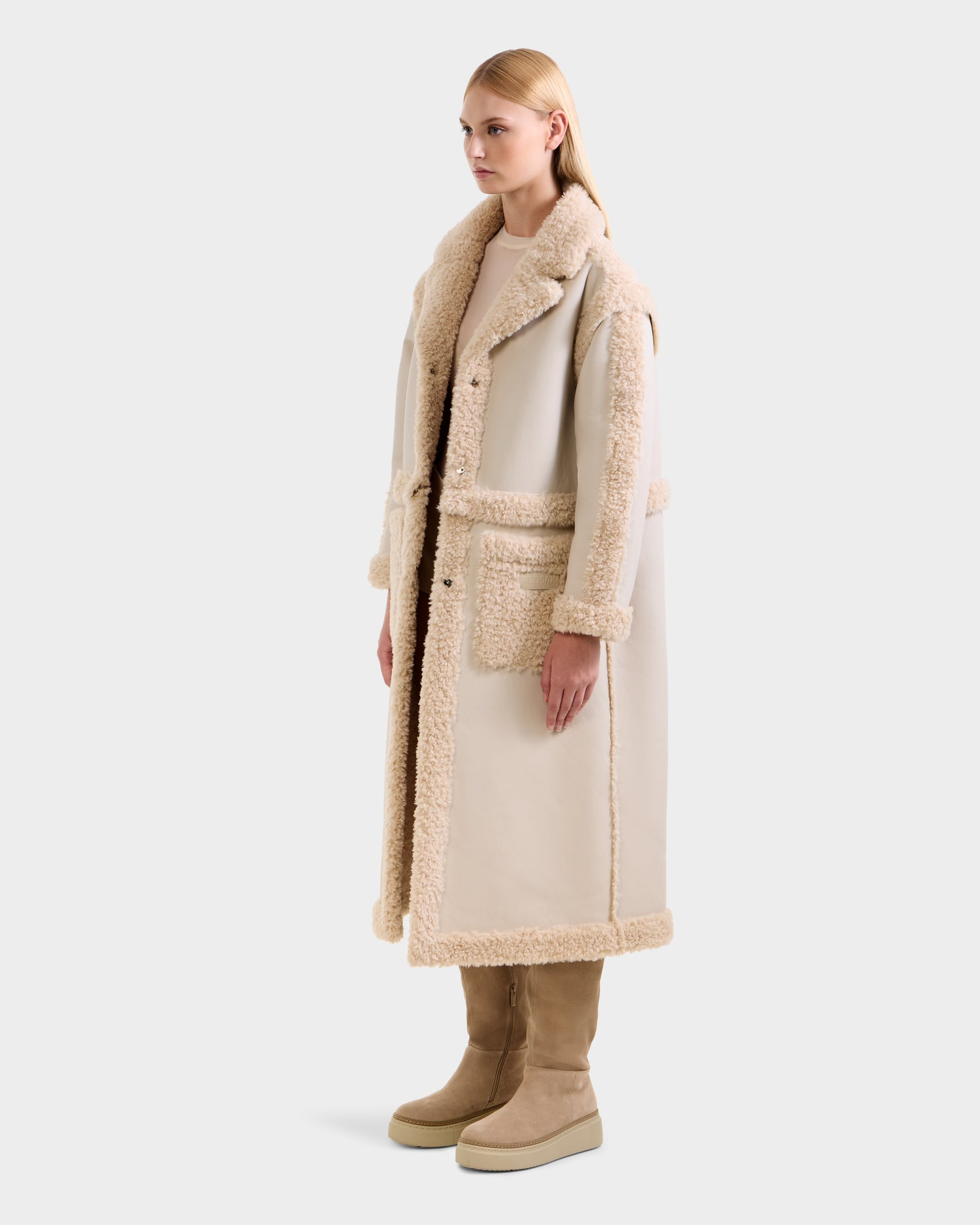 A woman wears the Rudsak PALOMA - 8125675 Vanilla long light beige faux shearling coat with tan fleece trim and large pockets, paired with matching tan boots, facing slightly left against a plain light background.