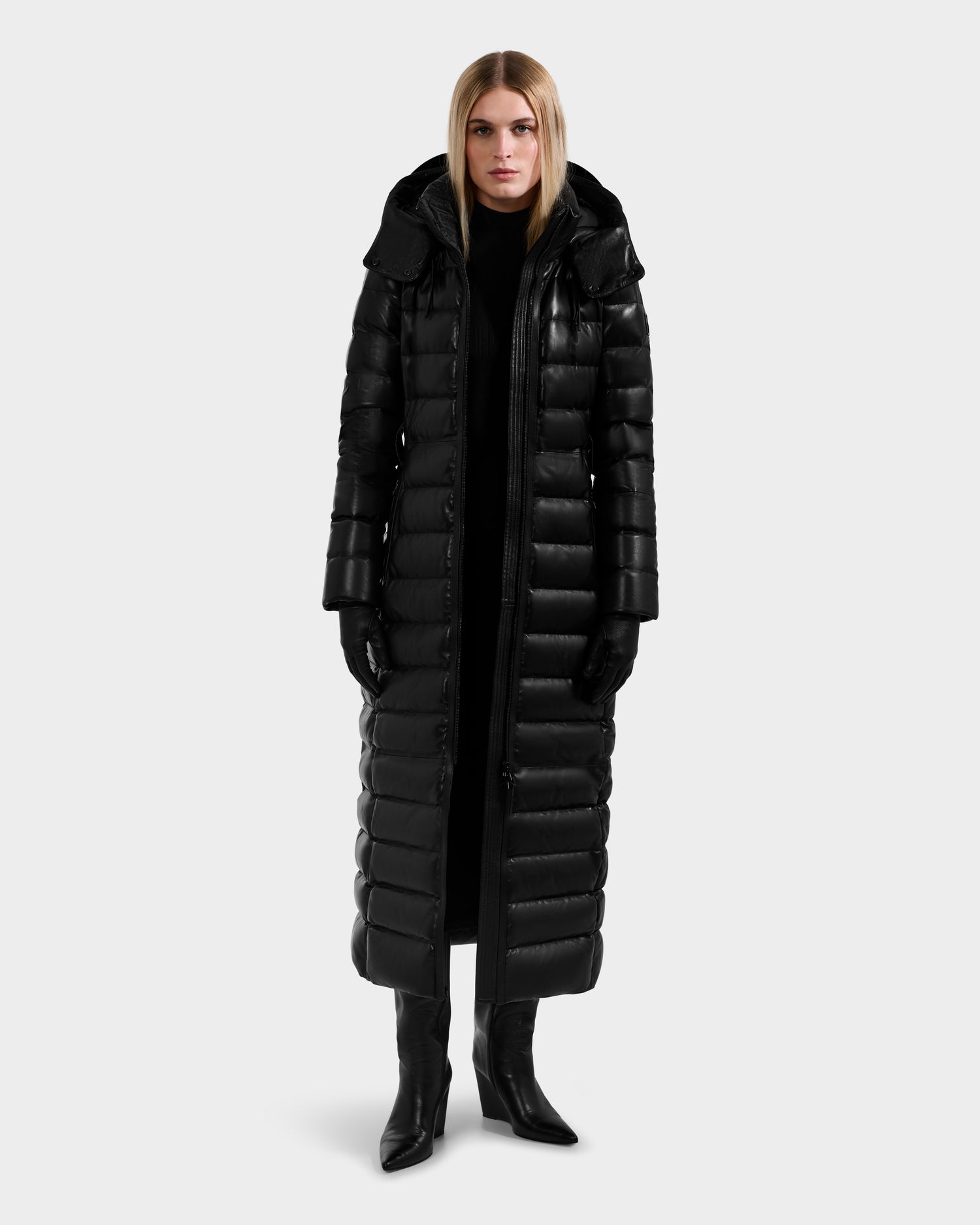 A person wears the Rudsak PELIN LX - 8125692 Black long leather down puffer women’s maxi coat with a hood, along with black gloves and boots, standing against a plain light gray background.