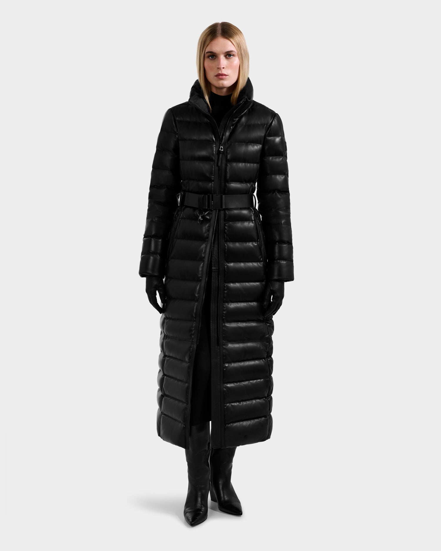 A woman faces forward wearing the Rudsak PELIN LX - 8125692 Black leather down maxi coat with a belt, paired with black gloves and heeled boots, set against a plain light grey background.