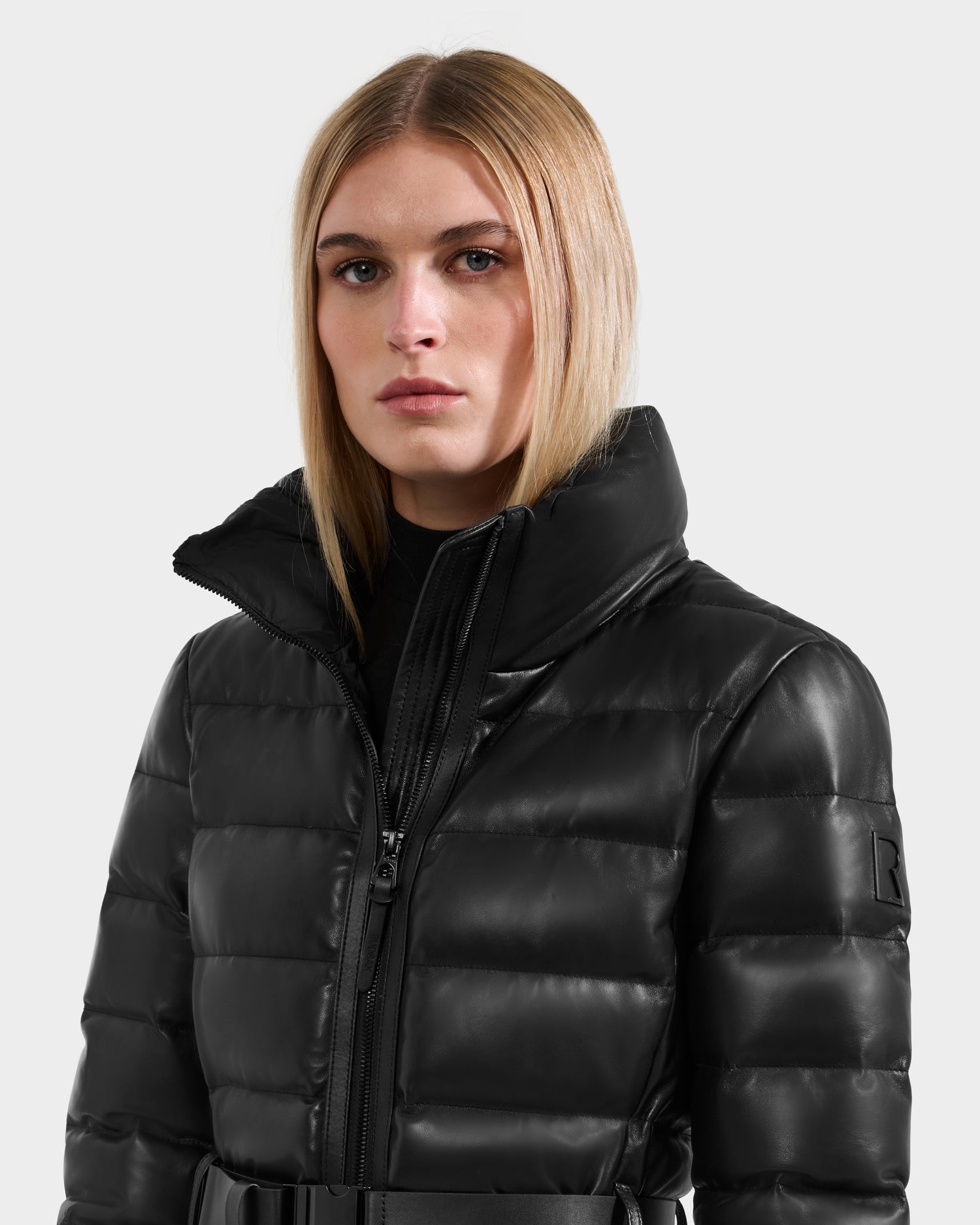 A person with straight blonde hair wears the Rudsak PELIN LX - 8125692 Black, a shiny black leather down puffer jacket with a high collar and zip-up front, standing against a plain light background.
