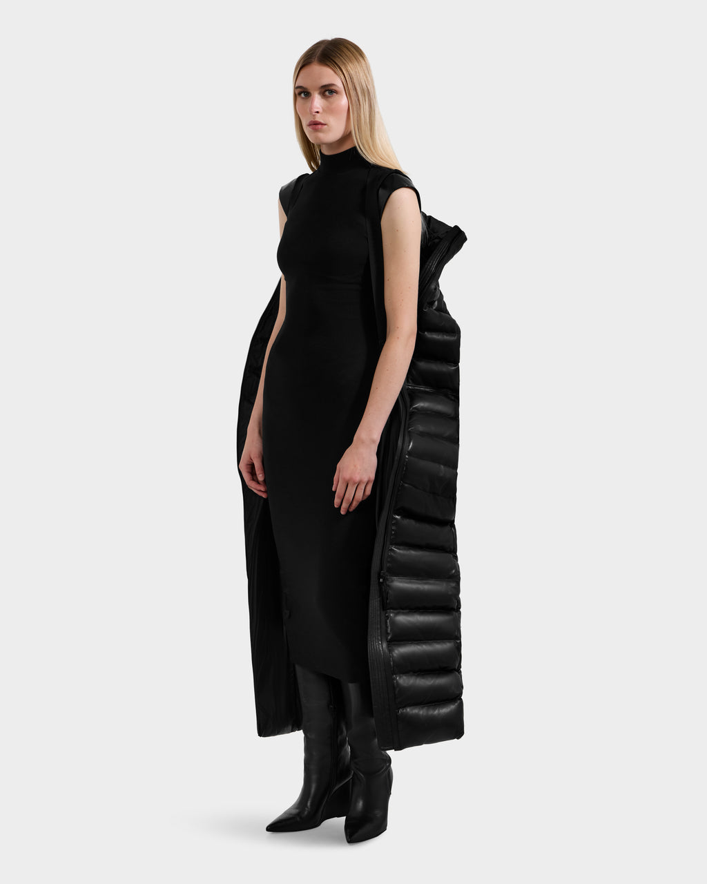 A woman with straight blonde hair wears a sleeveless black dress, tall black boots, and a Rudsak PELIN LX - 8125692 Black maxi coat styled as a leather down puffer draped over her shoulders, standing against a plain white background.