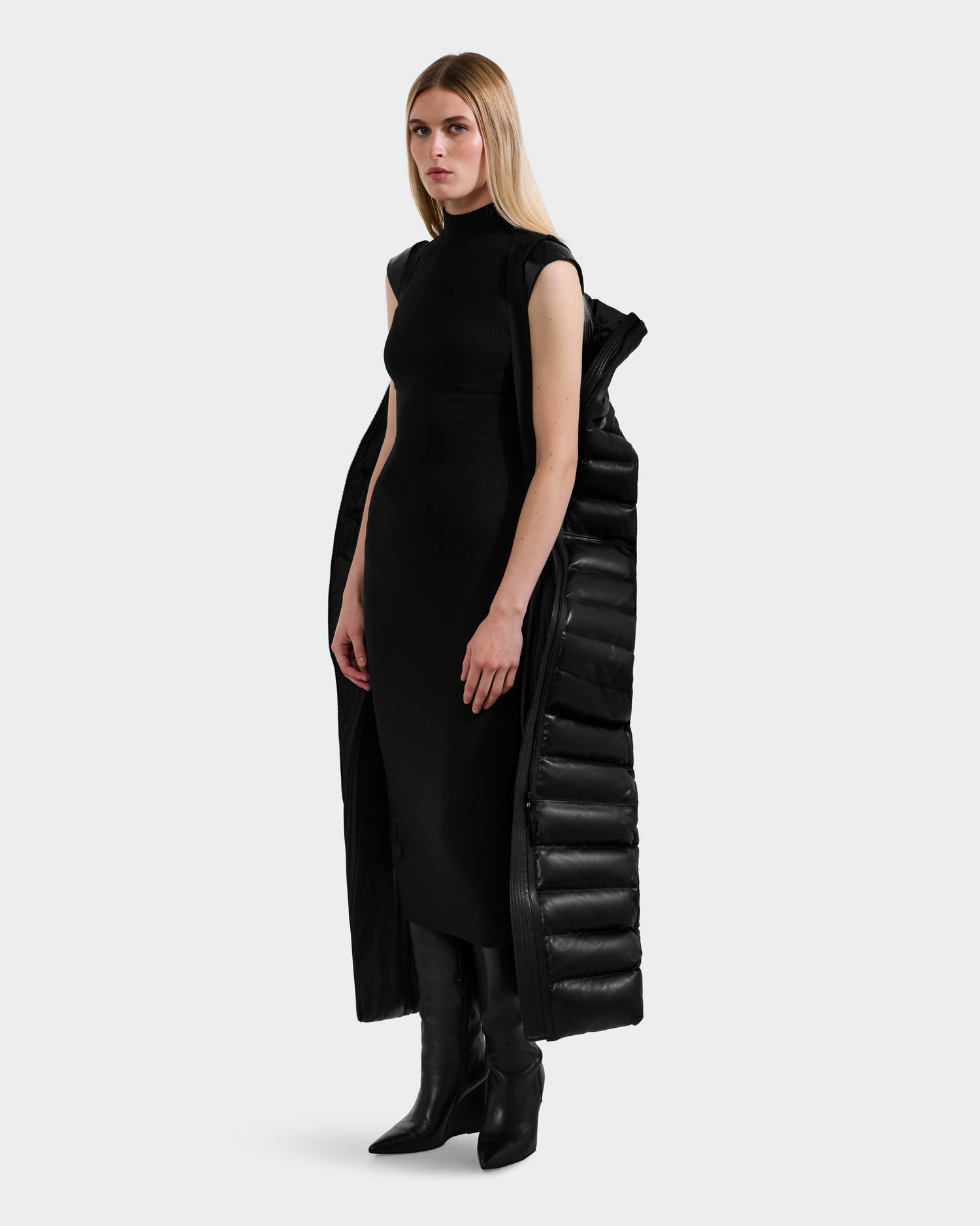 A woman with straight blonde hair wears a sleeveless black dress, tall black boots, and a Rudsak PELIN LX - 8125692 Black maxi coat styled as a leather down puffer draped over her shoulders, standing against a plain white background.
