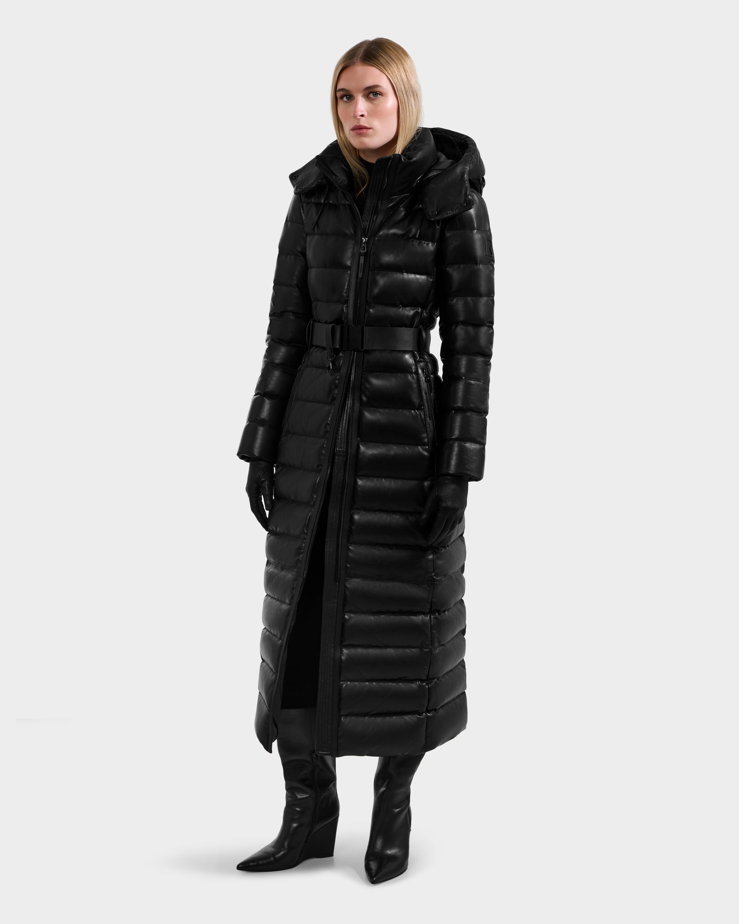 A woman in the Rudsak PELIN LX - 8125692 Black leather down maxi coat with a belt, black gloves, and heeled boots stands against a plain light background, gazing slightly to the side.
