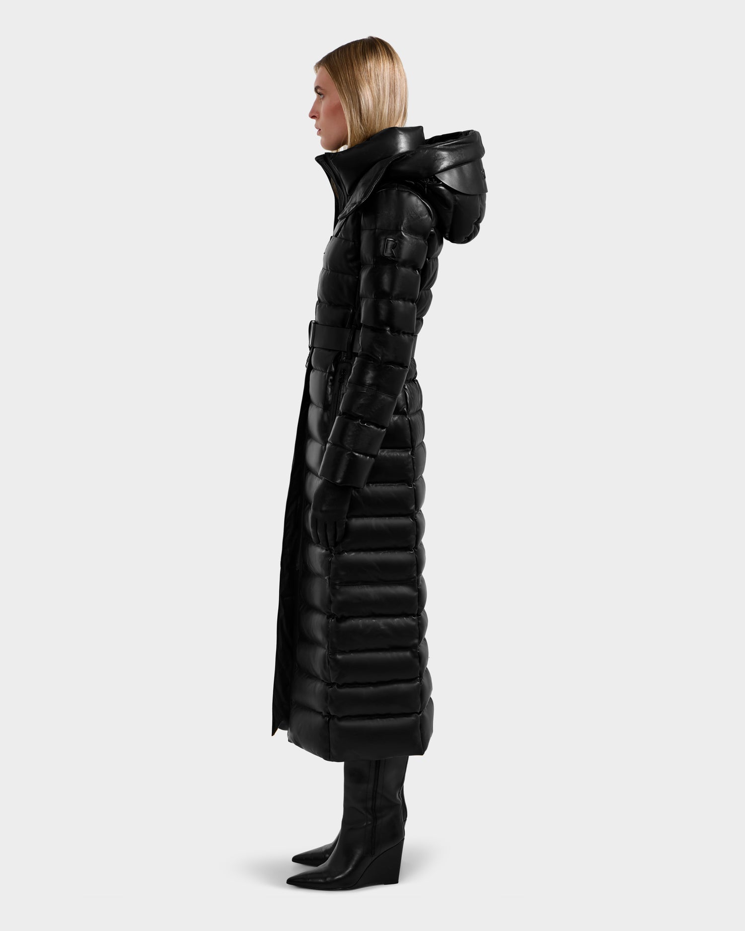 A woman stands in profile wearing the Rudsak PELIN LX - 8125692 Black women’s maxi coat with a hood, black gloves, and heeled boots against a plain light gray background.