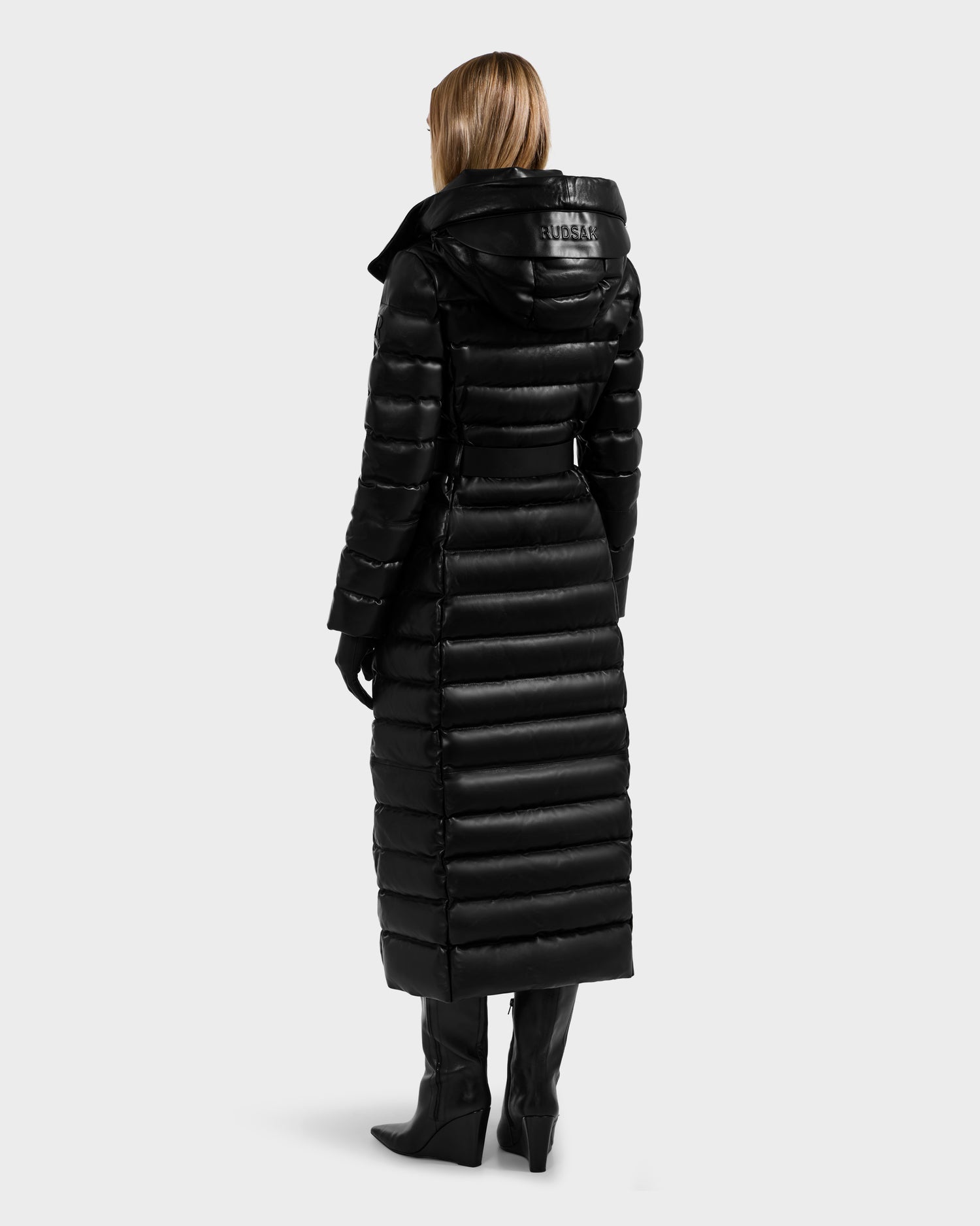 A person with blonde hair wears the Rudsak PELIN LX - 8125692 Black women’s maxi coat with a hood, paired with black gloves and heeled boots. They are standing, facing away, showing the outfit’s back against a plain light background.