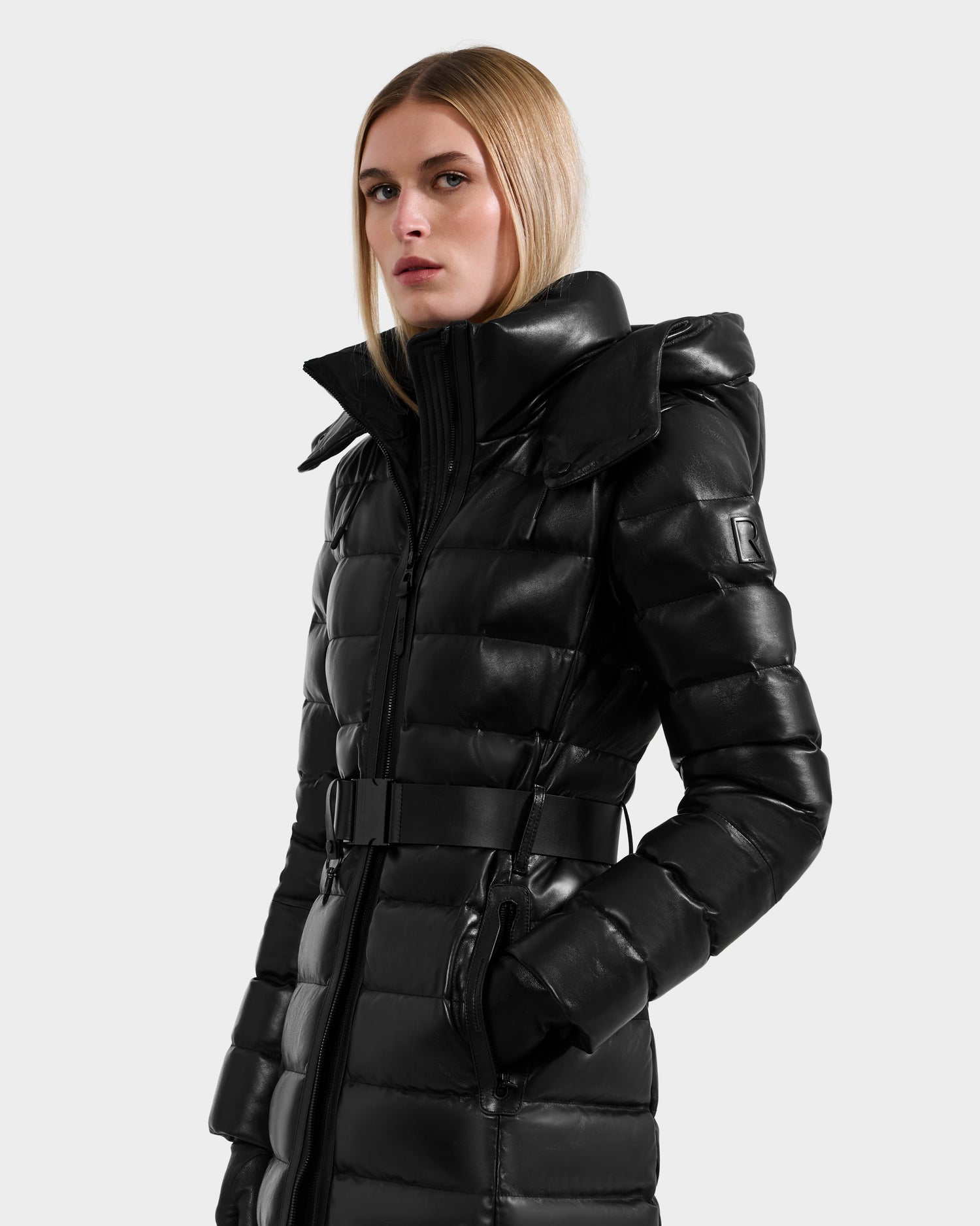 A woman with straight blonde hair wears the Rudsak PELIN LX - 8125692 Black leather down puffer coat with a high collar and belted waist, paired with gloves, standing against a plain light gray background.