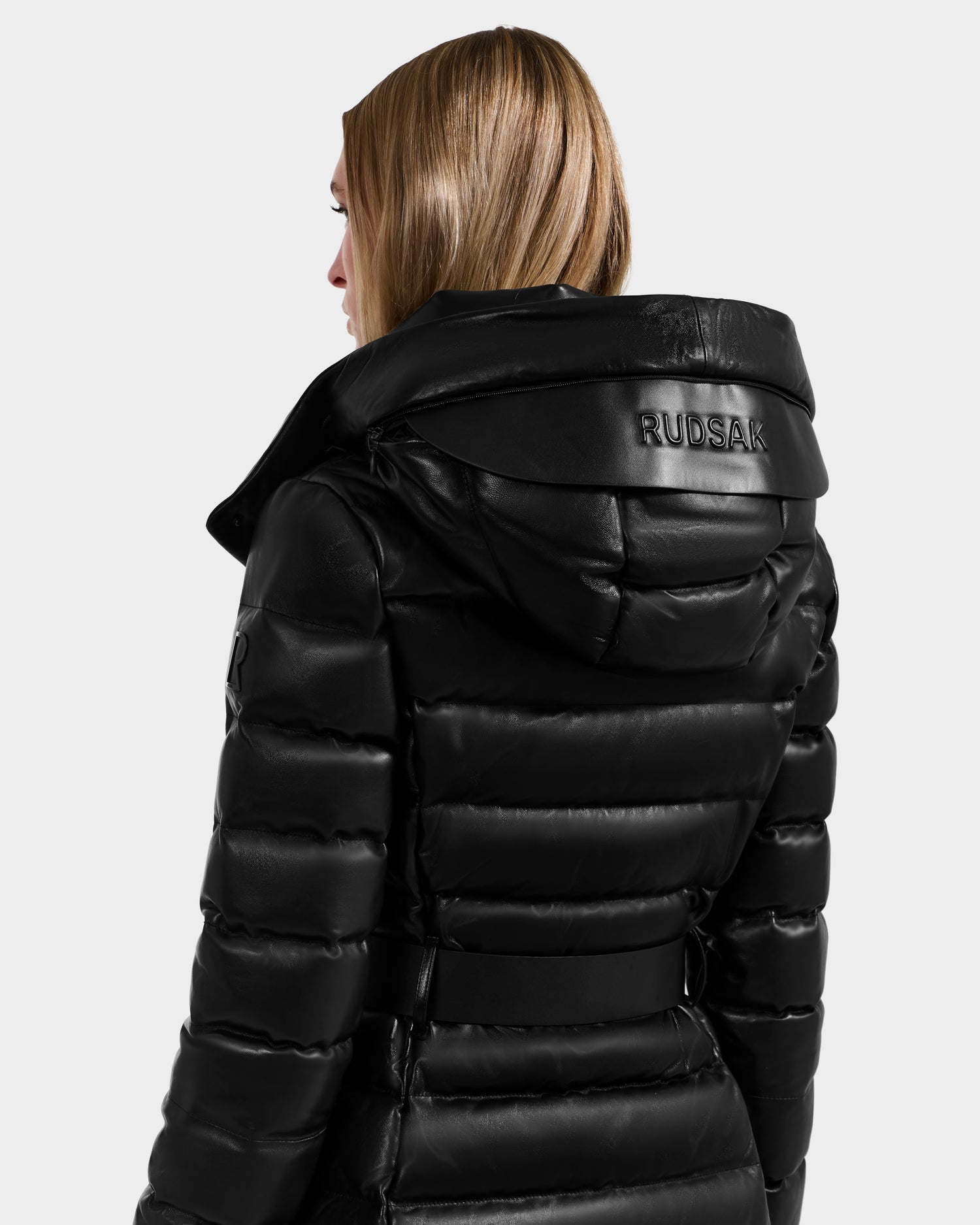 A person with straight, light brown hair wears a shiny black Rudsak PELIN LX - 8125692 women’s maxi coat with a hood. The leather down puffer has the brand name embossed on the hood and is shown from the back against a plain background.