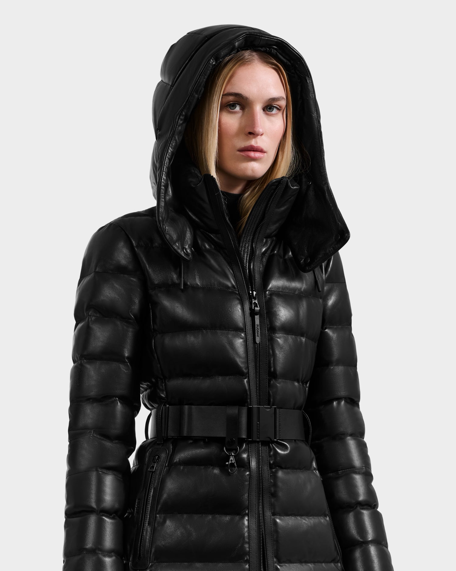 A woman stands against a light gray background wearing the Rudsak PELIN LX - 8125692 Black puffer coat, featuring a hood, quilted design, and belted waist.