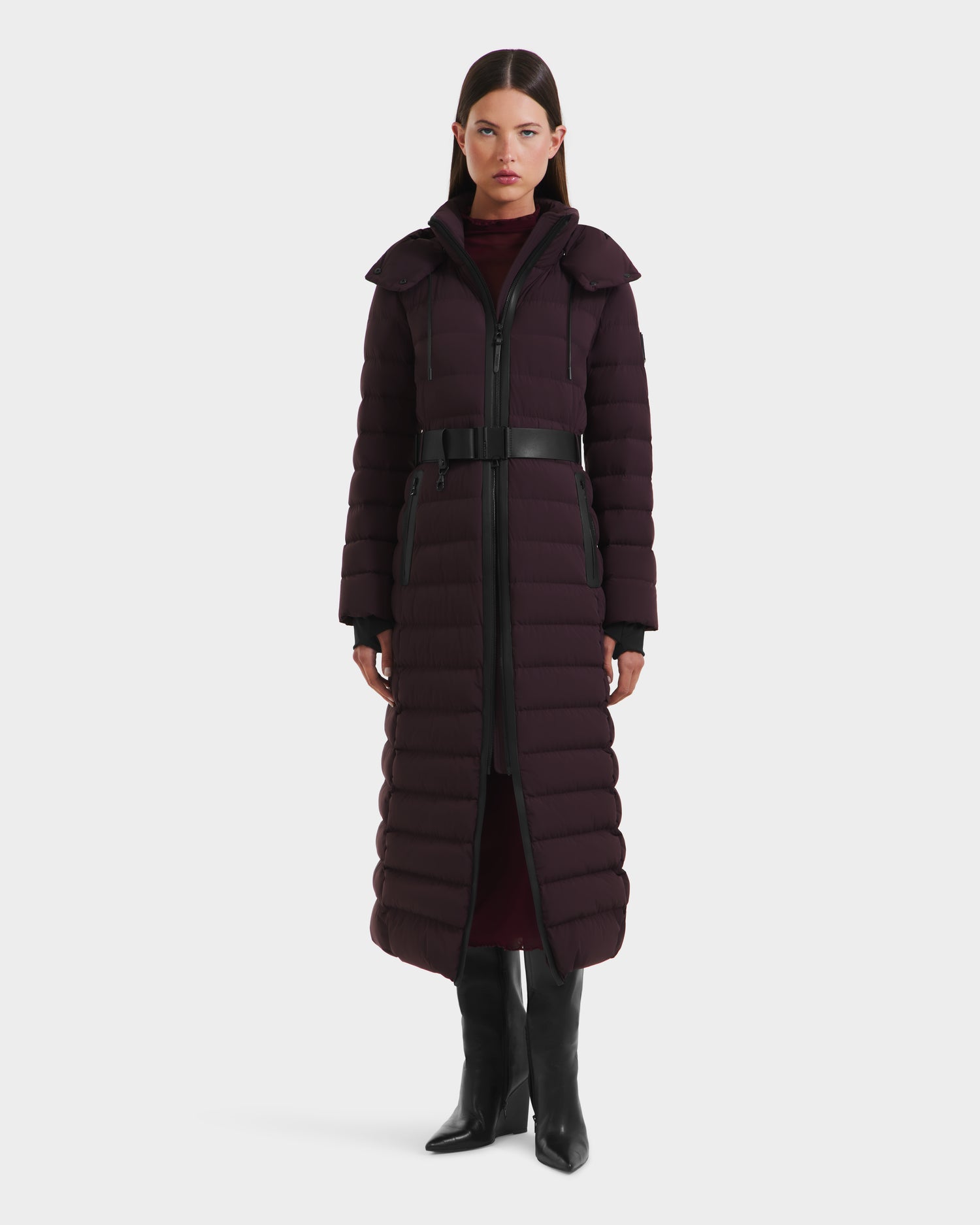 A woman faces forward in the Rudsak PELIN L - 8124815 Bordeaux, a long down quilted coat with a belted waist and high collar, paired with black knee-high boots, set against a plain white background.