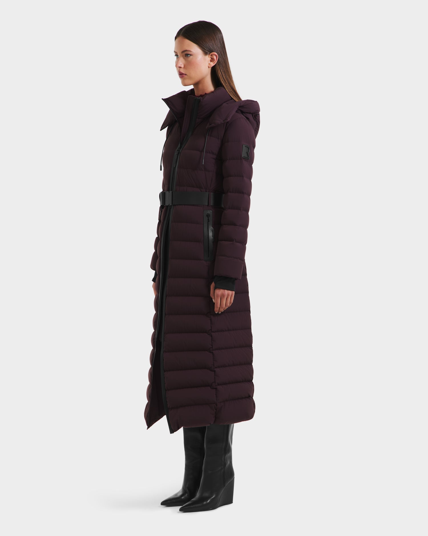A woman in profile wears the Rudsak PELIN L - 8124815 Bordeaux, a long dark purple RDS-certified down quilted winter coat with a hood and black belt, paired with black knee-high boots. The coat extends nearly to her ankles. Plain white background.