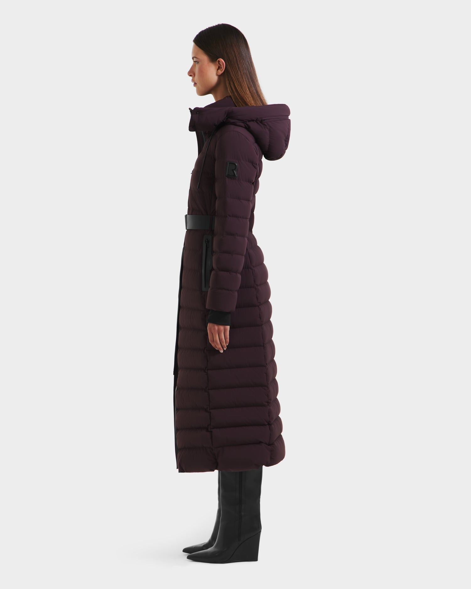 A woman in profile models the Rudsak PELIN L - 8124815 Bordeaux long quilted hooded winter coat, belted at the waist, with straight brown hair and black knee-high boots against a plain white background.