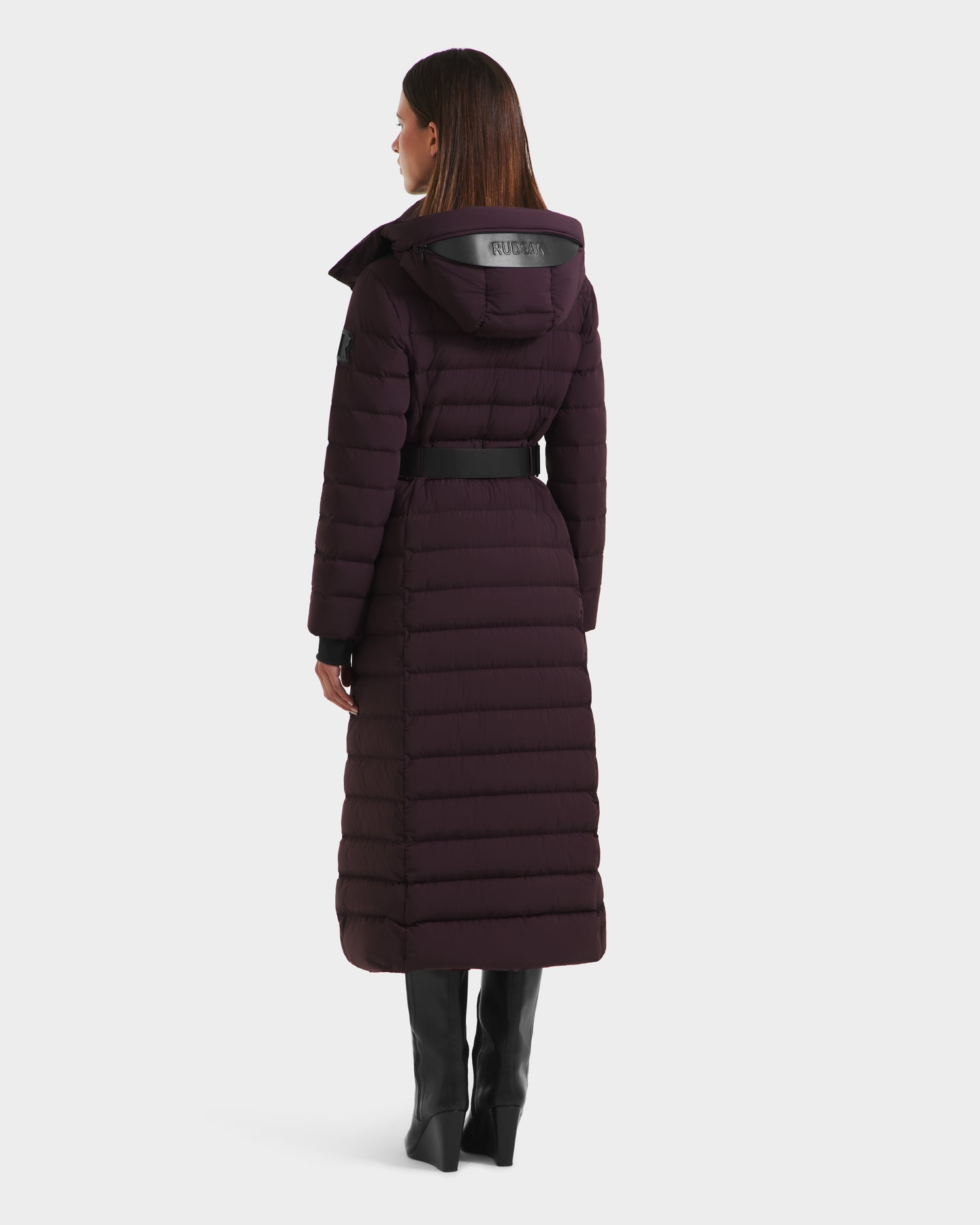A woman faces away, wearing the Rudsak PELIN L - 8124815 Bordeaux down puffer coat with a hooded belt design and horizontal quilting, paired with black knee-high boots. The long coat extends to mid-calf for winter warmth and style.