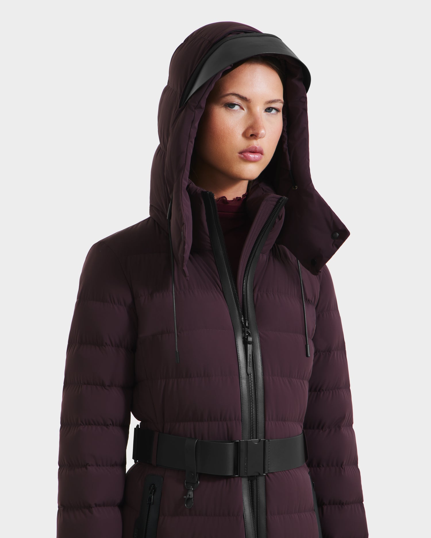 A woman models the Rudsak PELIN L - 8124815 Bordeaux, a dark purple, RDS-certified down winter coat with a belt, hood, and black zipper, against a plain white background.