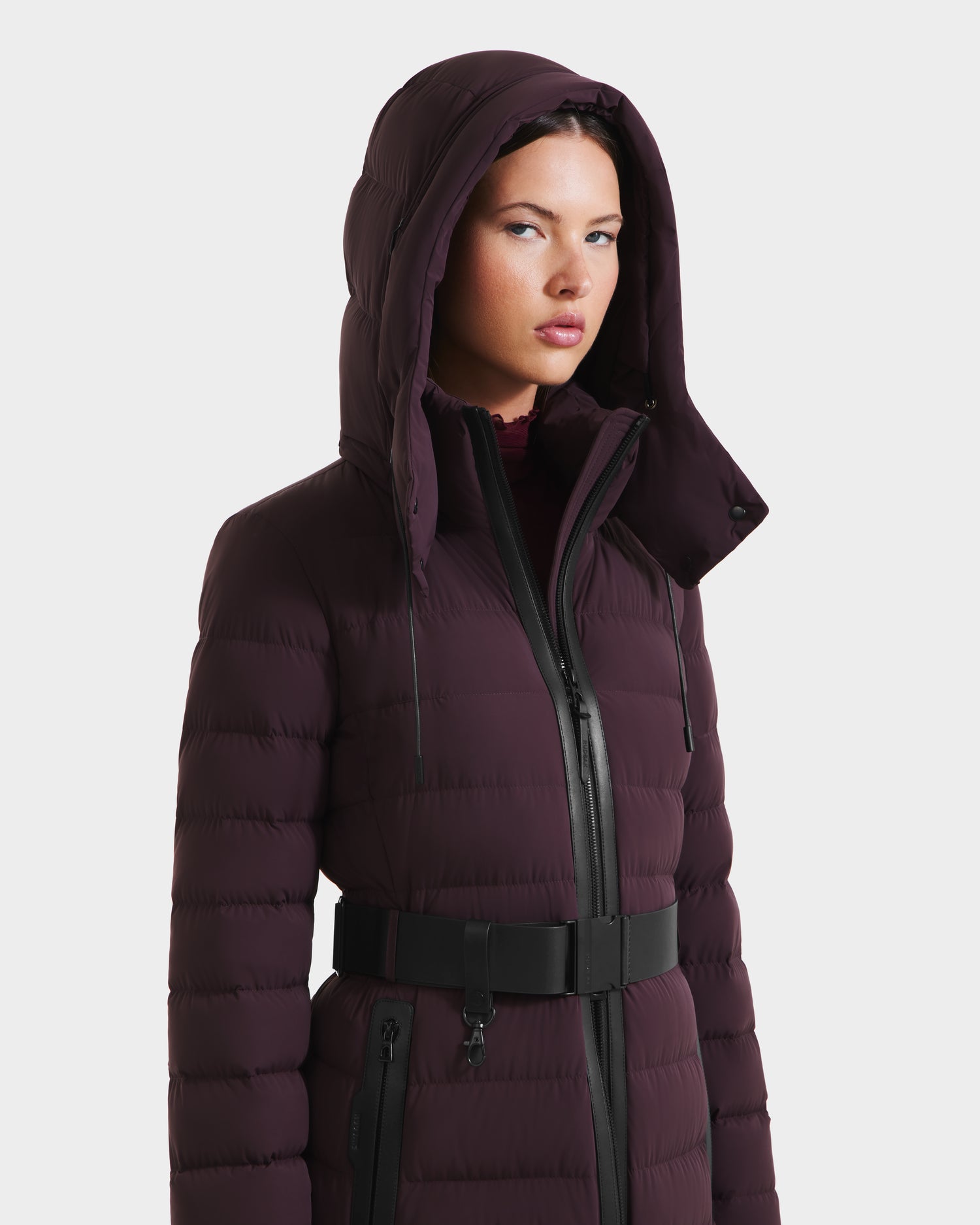 A woman models the Rudsak PELIN L - 8124815 Bordeaux down puffer coat, featuring a hood and belt, against a plain white background with a neutral expression.