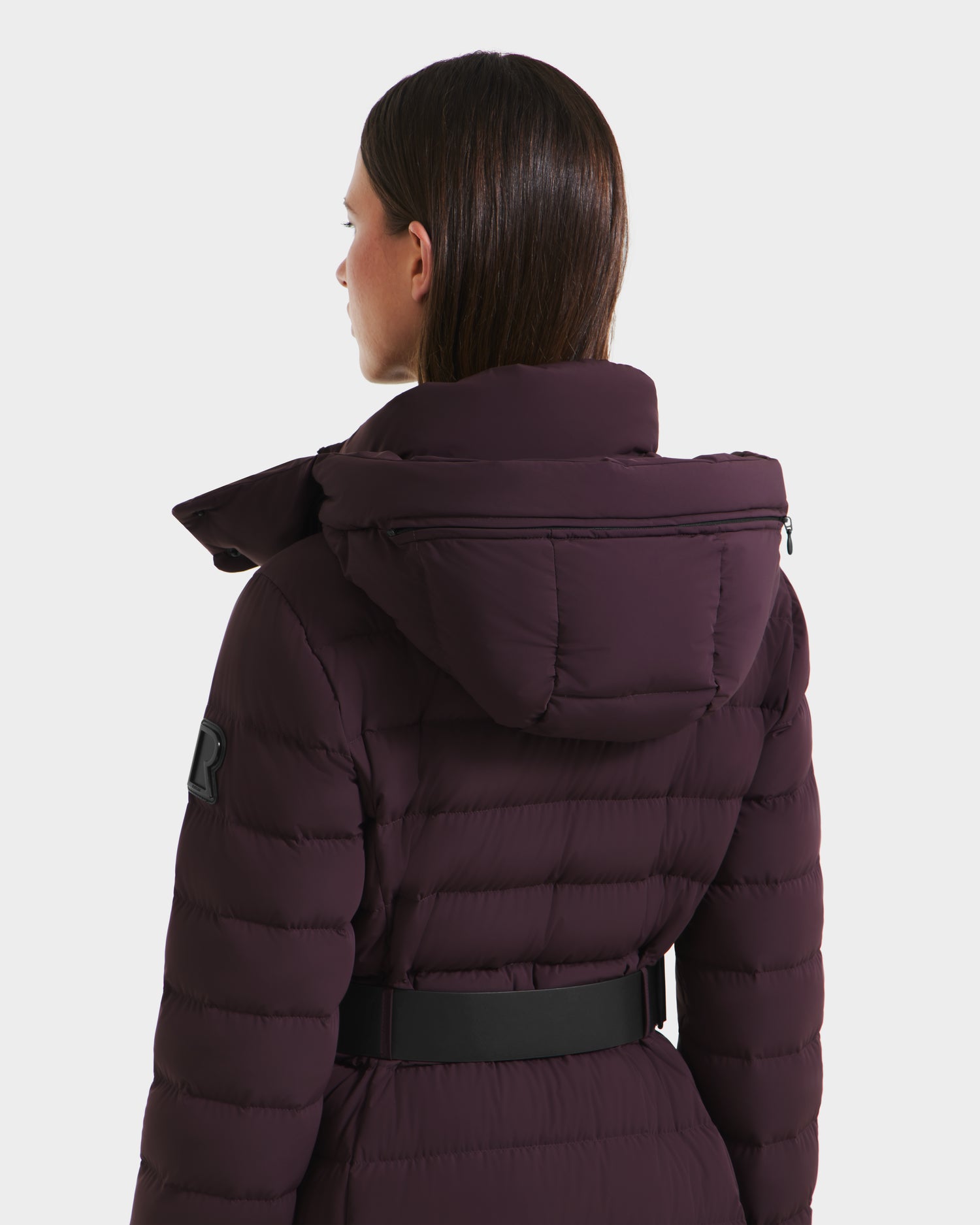A woman with straight brown hair is seen from behind wearing the Rudsak PELIN L - 8124815 Bordeaux quilted hooded winter coat with a black belt, set against a plain light background.