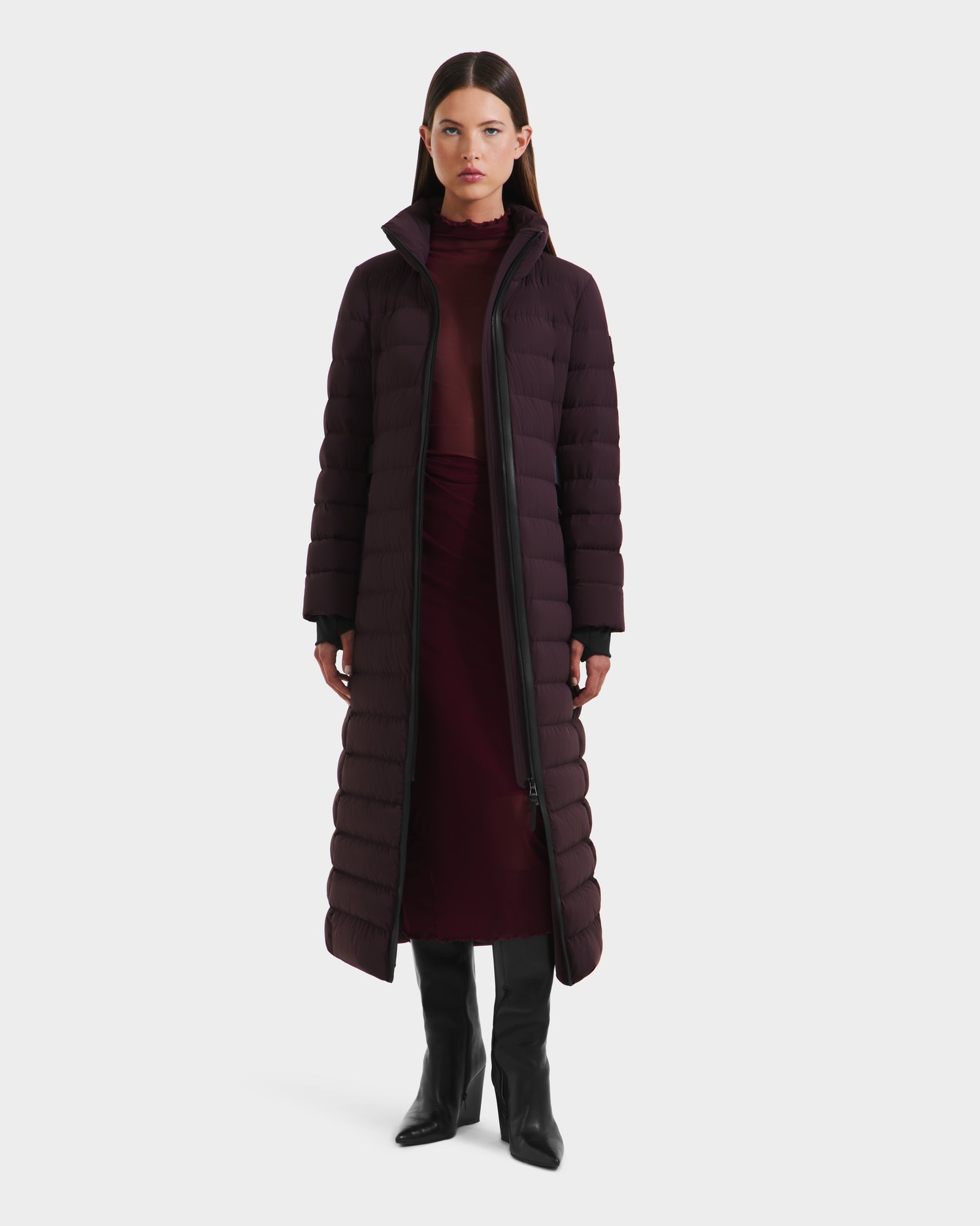 A woman faces forward in a Rudsak PELIN L - 8124815 Bordeaux long down puffer coat over a burgundy dress and black knee-high boots, standing against a plain white background.