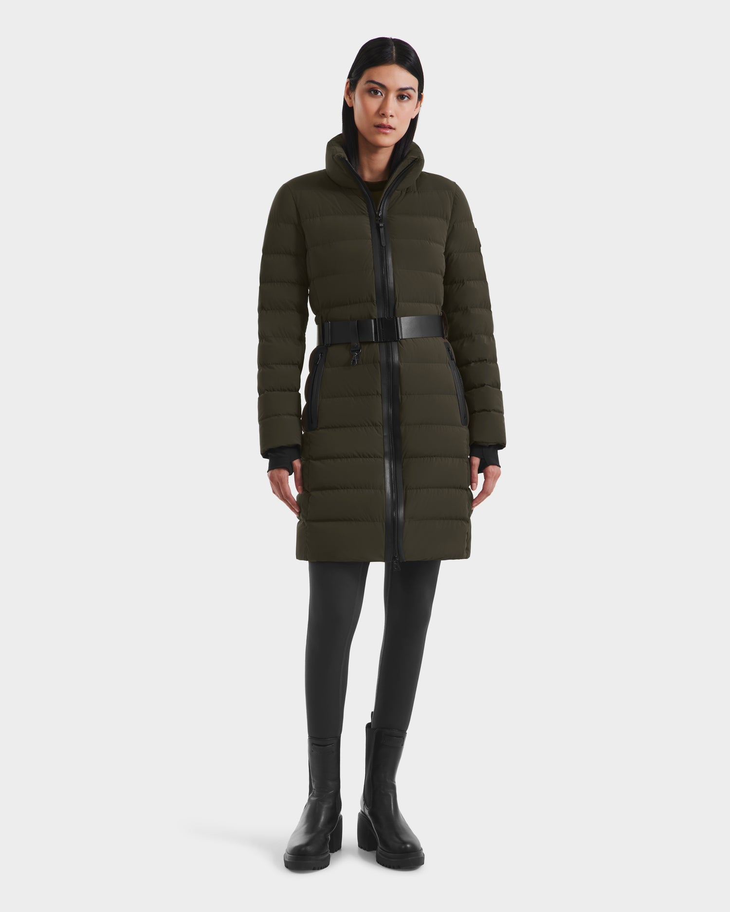 A woman wearing the Rudsak PELIN M - 8124814 Dark Olive RDS-certified knee-length quilted winter jacket with a high collar, black belt, black leggings, and black ankle boots stands against a plain white background.