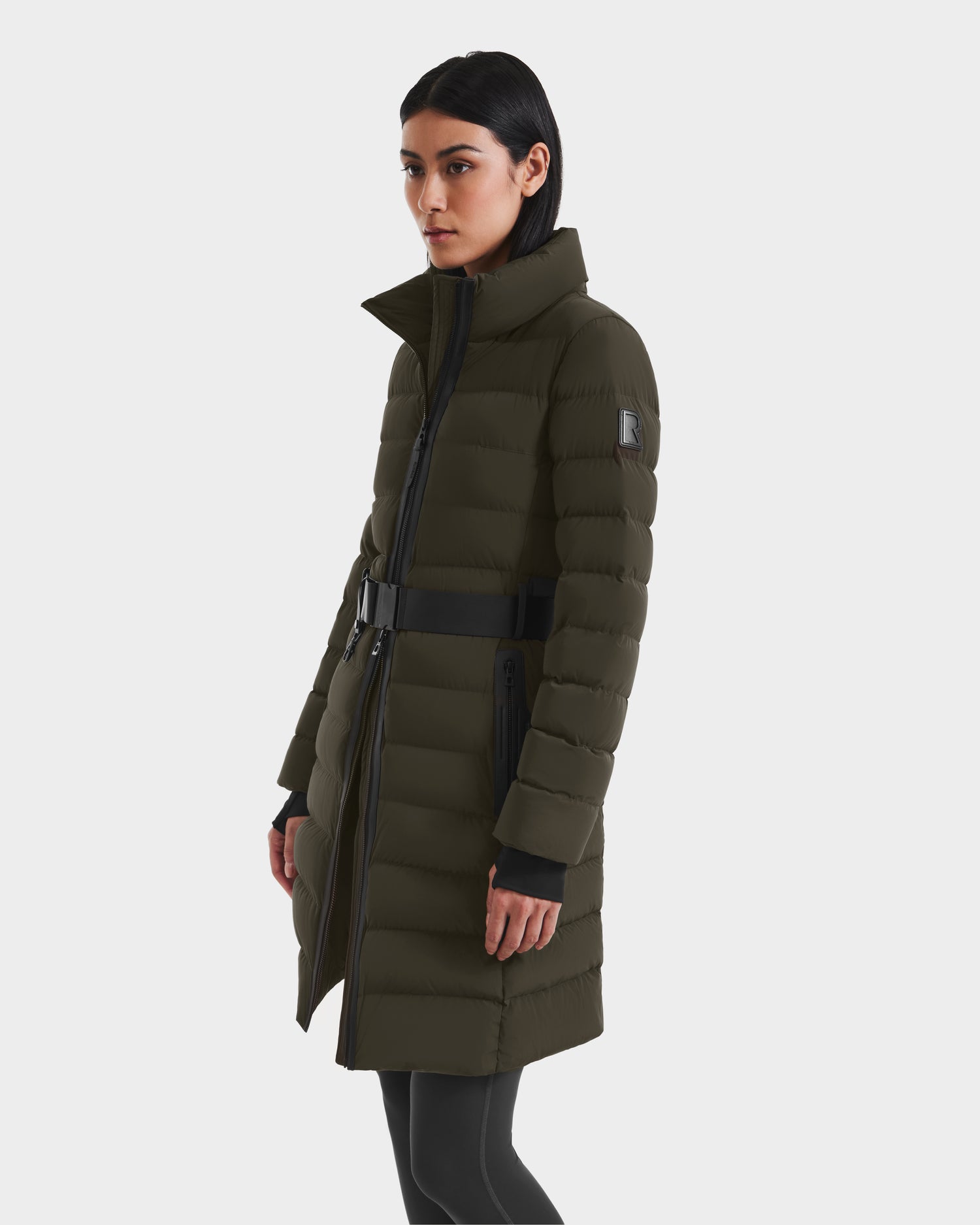 A woman wearing the Rudsak PELIN M - 8124814 Dark Olive down puffer with a high collar, black belt, and zippered pockets stands against a plain light background.