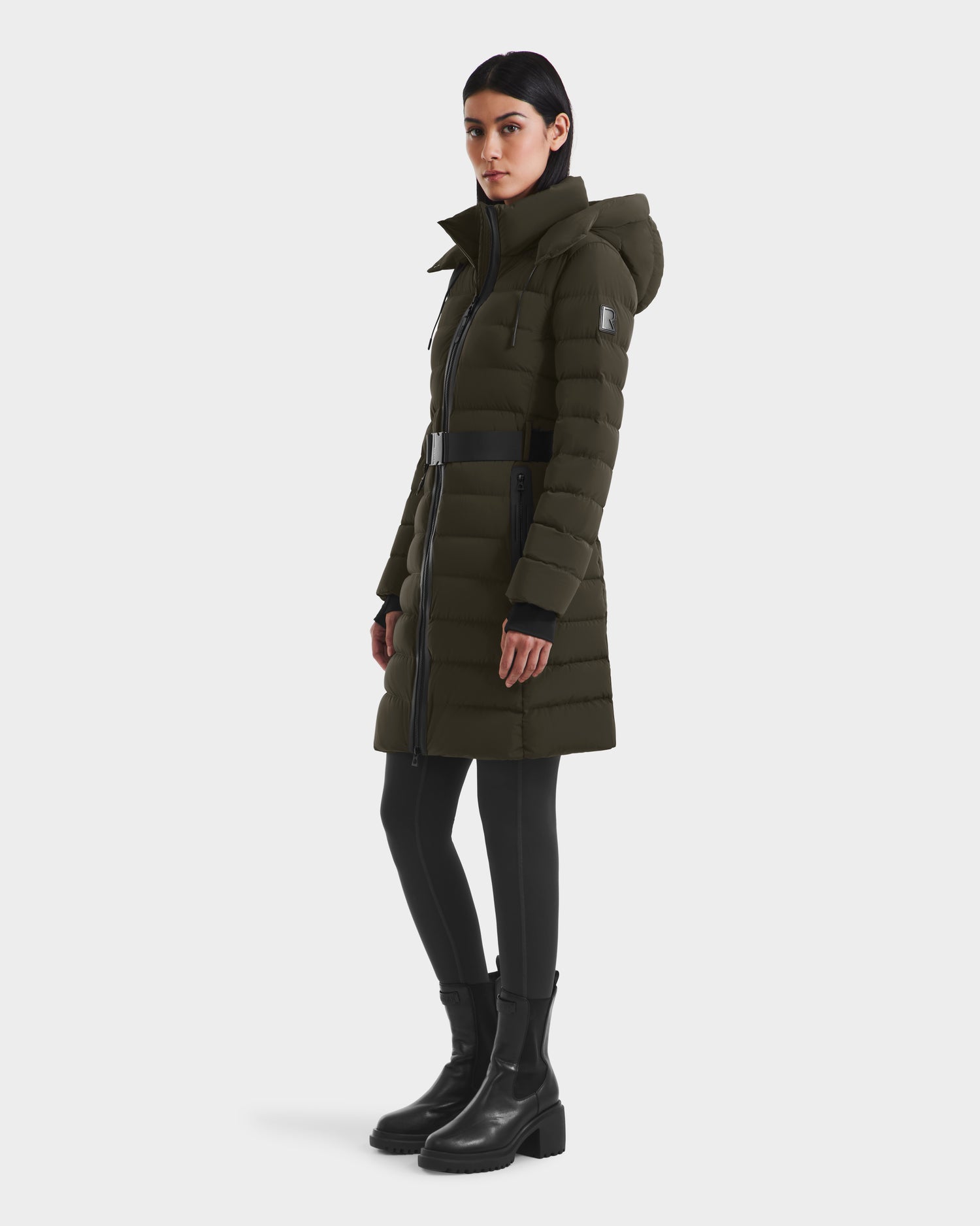 A woman wearing the Rudsak PELIN M - 8124814 Dark Olive RDS-certified down puffer with a hood, black leggings, and black ankle boots stands facing slightly to the side against a plain white background.
