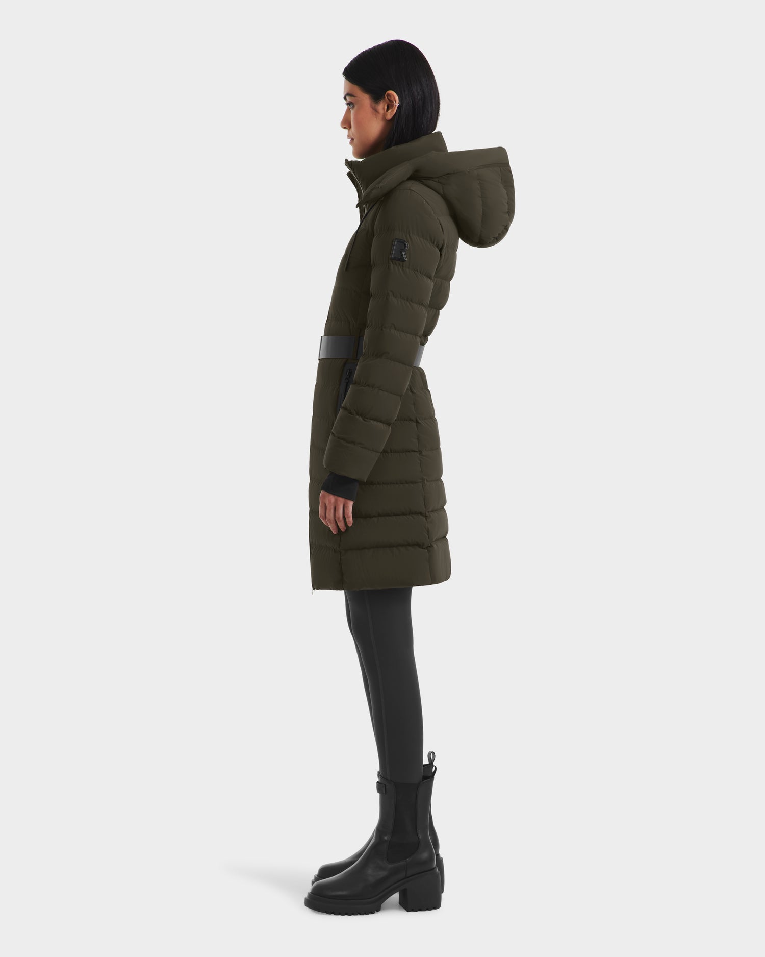 A person stands in profile wearing the Rudsak PELIN M - 8124814 Dark Olive, an RDS-certified women’s down puffer with a hood, paired with black leggings and black ankle boots against a plain white background.