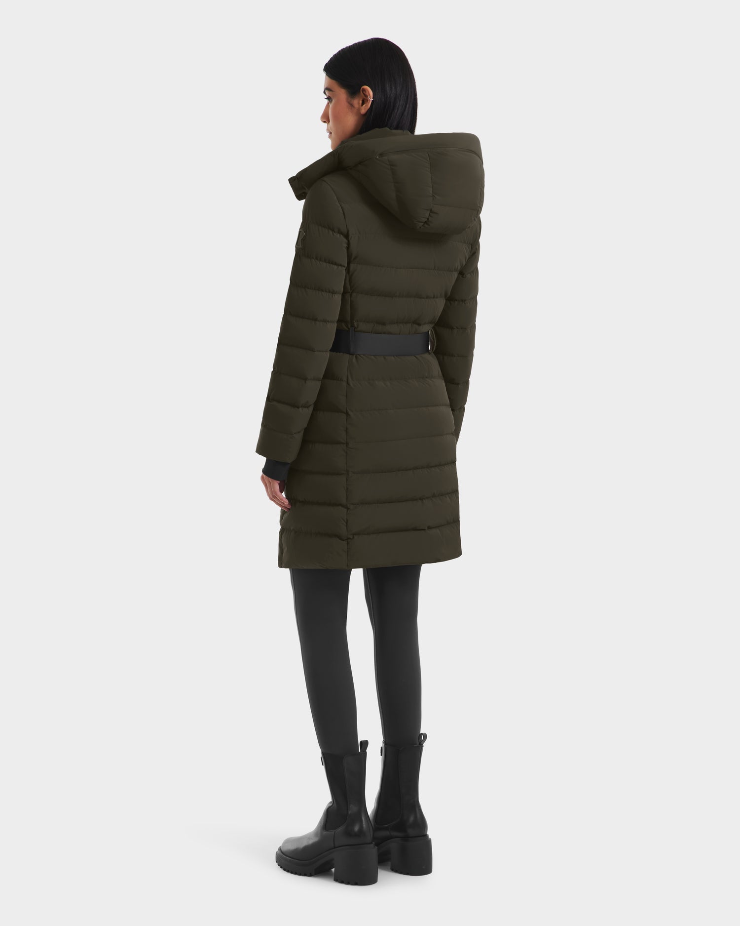 A woman with straight dark hair faces away, wearing the Rudsak PELIN M - 8124814 Dark Olive RDS-certified quilted winter jacket with a hood, black leggings, and black ankle boots.