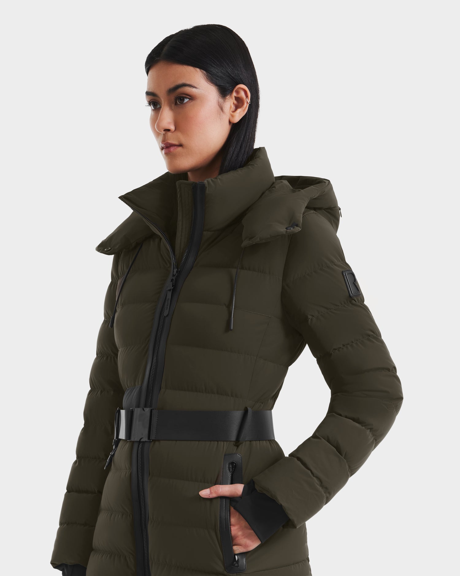 A woman with long dark hair models the Rudsak PELIN M - 8124814 Dark Olive belted women's down puffer coat with a high collar and zippered pockets, standing against a plain white background.
