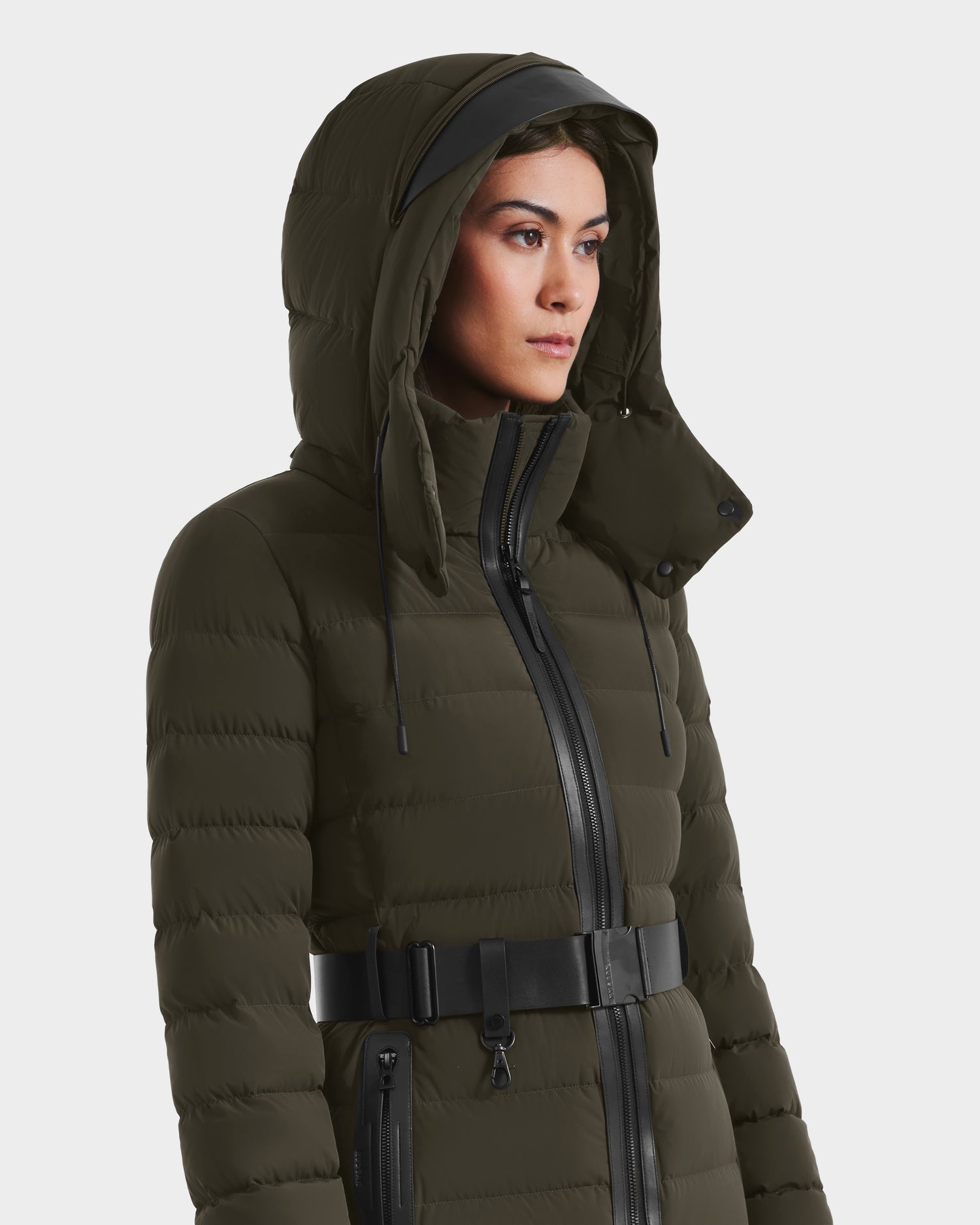 A woman models the Rudsak PELIN M - 8124814 Dark Olive jacket, featuring a front zipper, black belt, and high collar, standing against a plain white background.