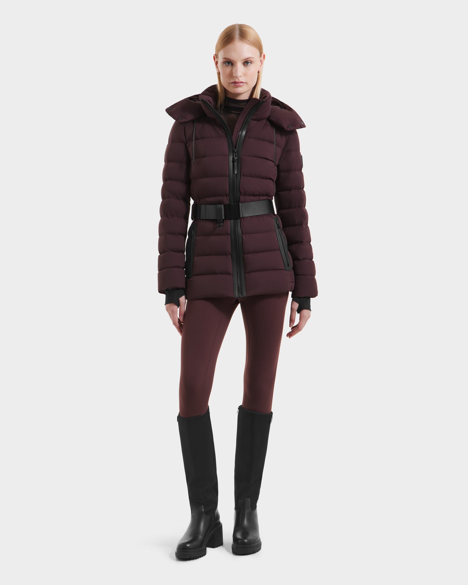 A woman in the Rudsak PELIN S - 8124813 Bordeaux quilted coat stands facing forward, paired with burgundy leggings and black knee-high boots, against a plain white background.