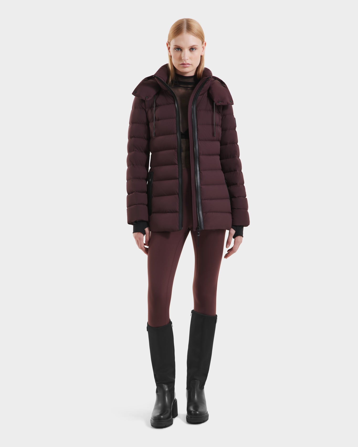 A woman faces forward against a plain white background, wearing the Rudsak PELIN S - 8124813 Bordeaux women’s down puffer jacket, paired with matching leggings, a sheer black top, and black knee-high boots.