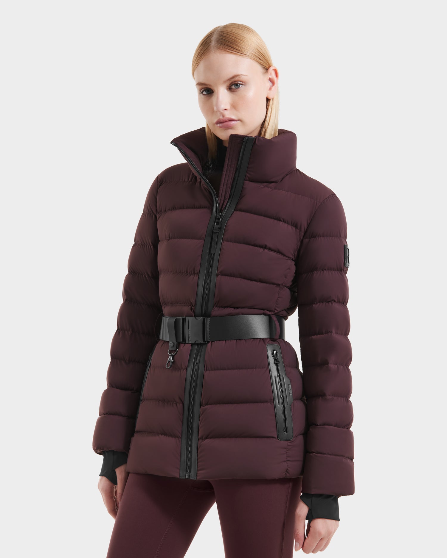 A woman wears the Rudsak PELIN S - 8124813 down puffer jacket in Bordeaux, featuring a black belt and zipper details, as she poses against a plain light background with a neutral expression.