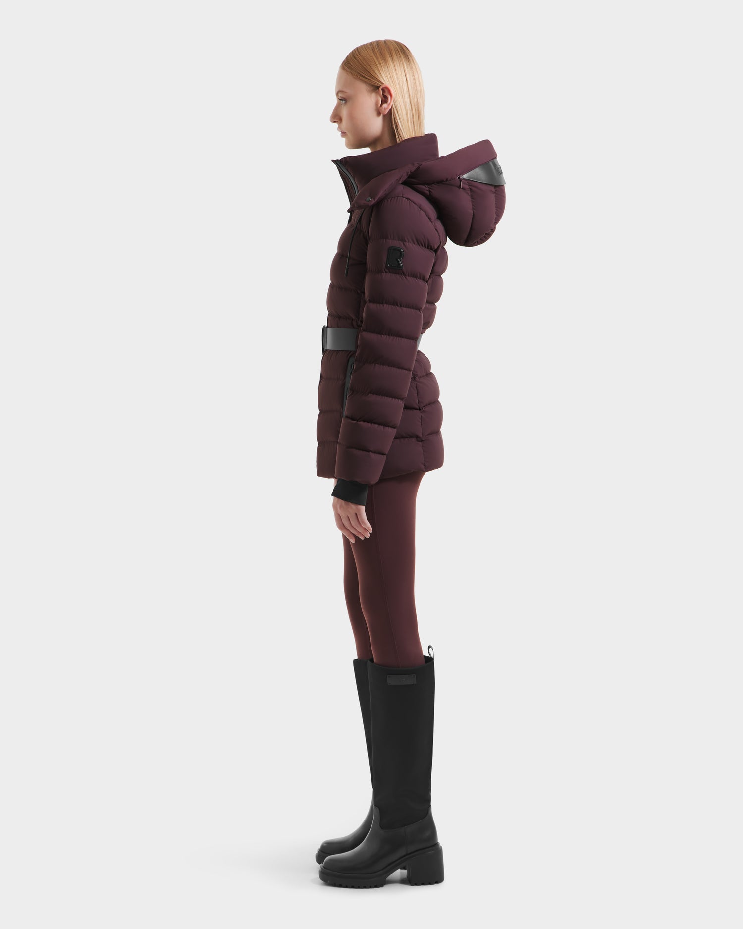 A person stands in profile wearing the Rudsak PELIN S - 8124813 Bordeaux womens down puffer with a large hood, matching leggings, and black knee-high boots against a plain white background.