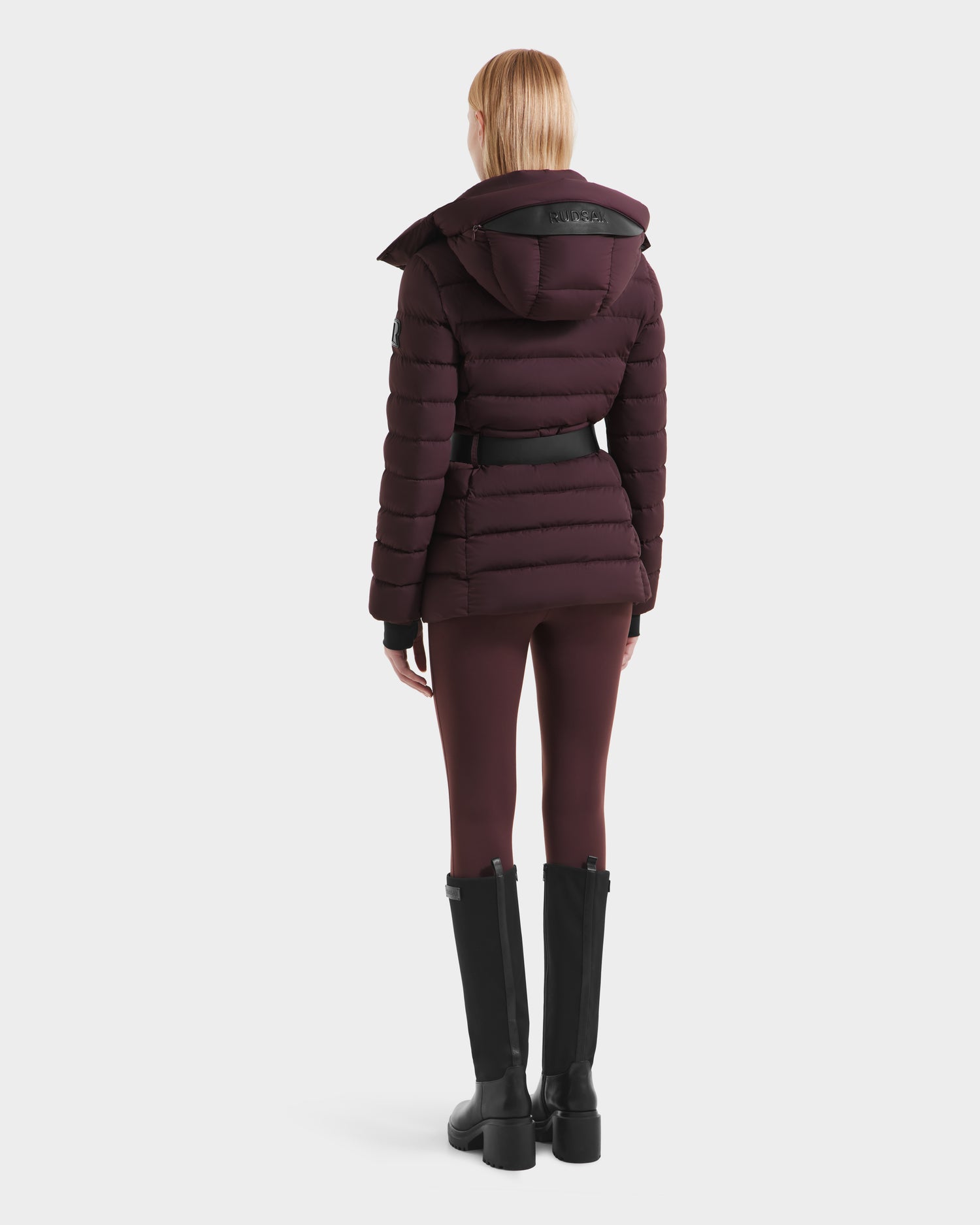 A woman with straight, shoulder-length blonde hair is seen from behind wearing the Rudsak PELIN S - 8124813 Bordeaux down puffer with hood, matching leggings, and tall black boots against a plain white background.