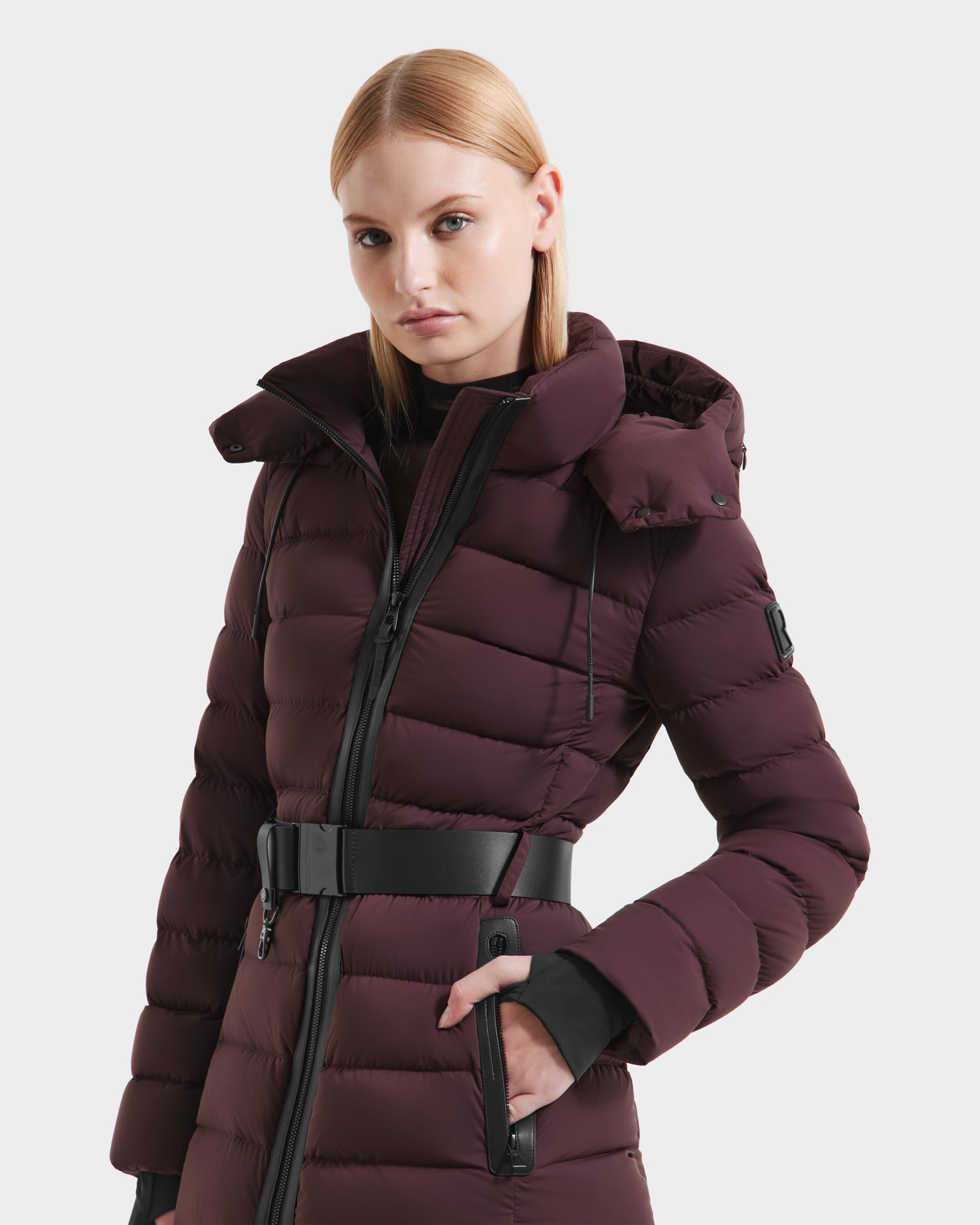 A woman with straight blonde hair models the Rudsak PELIN S - 8124813 Bordeaux, a belted dark burgundy quilted winter coat with a hood and high collar, posing with one hand in her pocket against a plain light background.