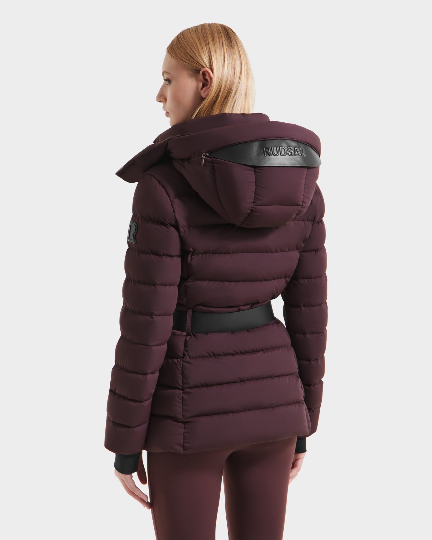 A person with straight, light brown hair is seen from the back wearing the Rudsak PELIN S - 8124813 Bordeaux womens down puffer, featuring RDS-certified down, a leather KUDSAI hood detail, black belt, sleeve patch, and quilted stitching.