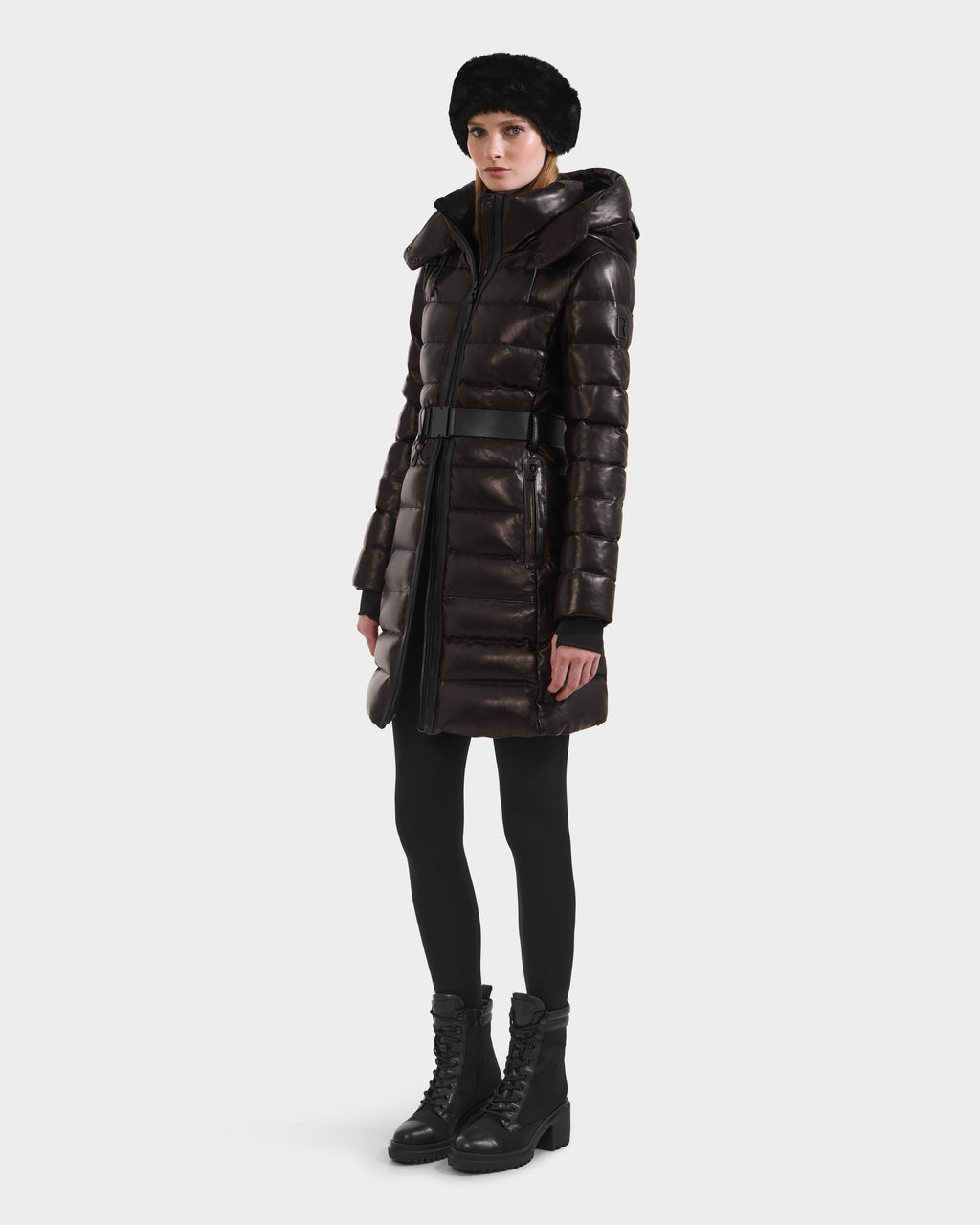 A person wearing the Rudsak PELIN X - 8125575 Black knee-length leather down puffer with a belted waist, black leggings, lace-up boots, and a black fur hat stands against a plain white background.