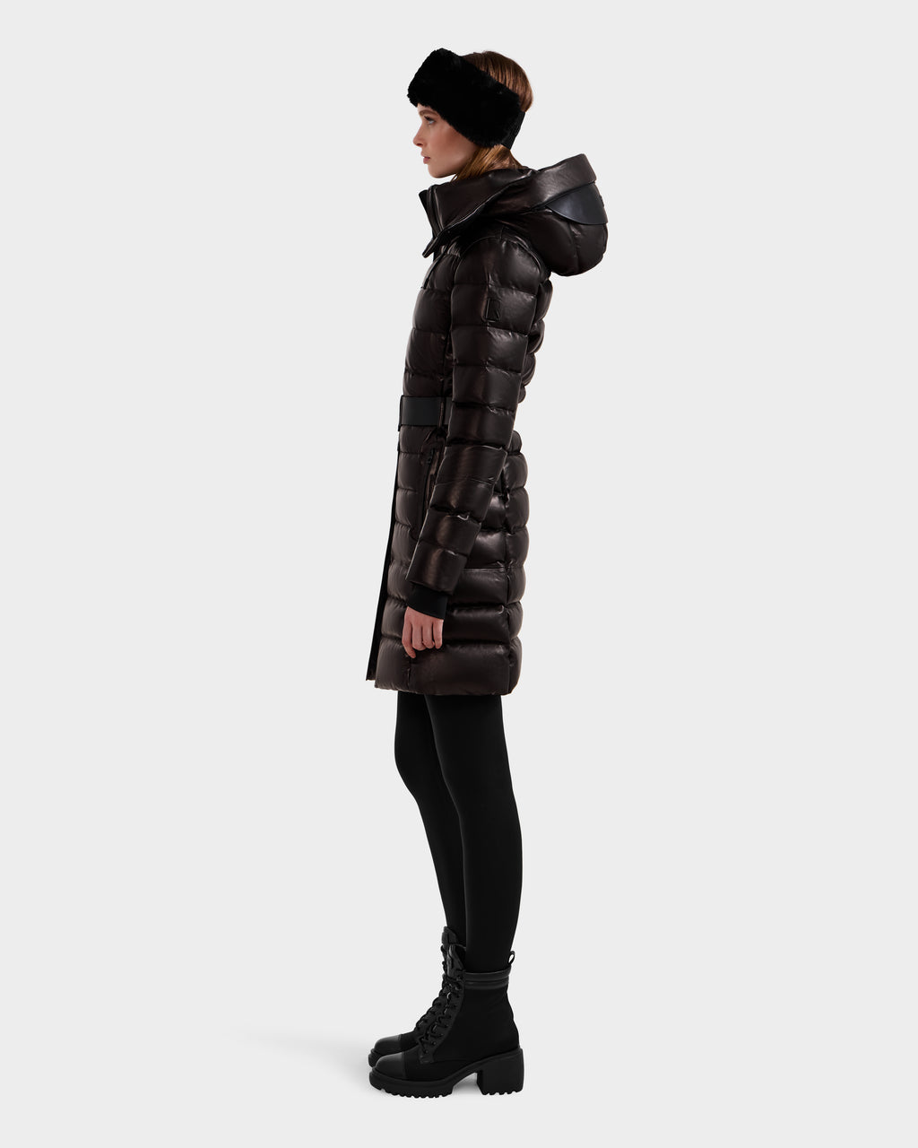 A person stands in profile wearing the Rudsak PELIN X - 8125575 Black women's leather down puffer winter coat with a hood, paired with black leggings, lace-up boots, and a black headband against a plain light background.