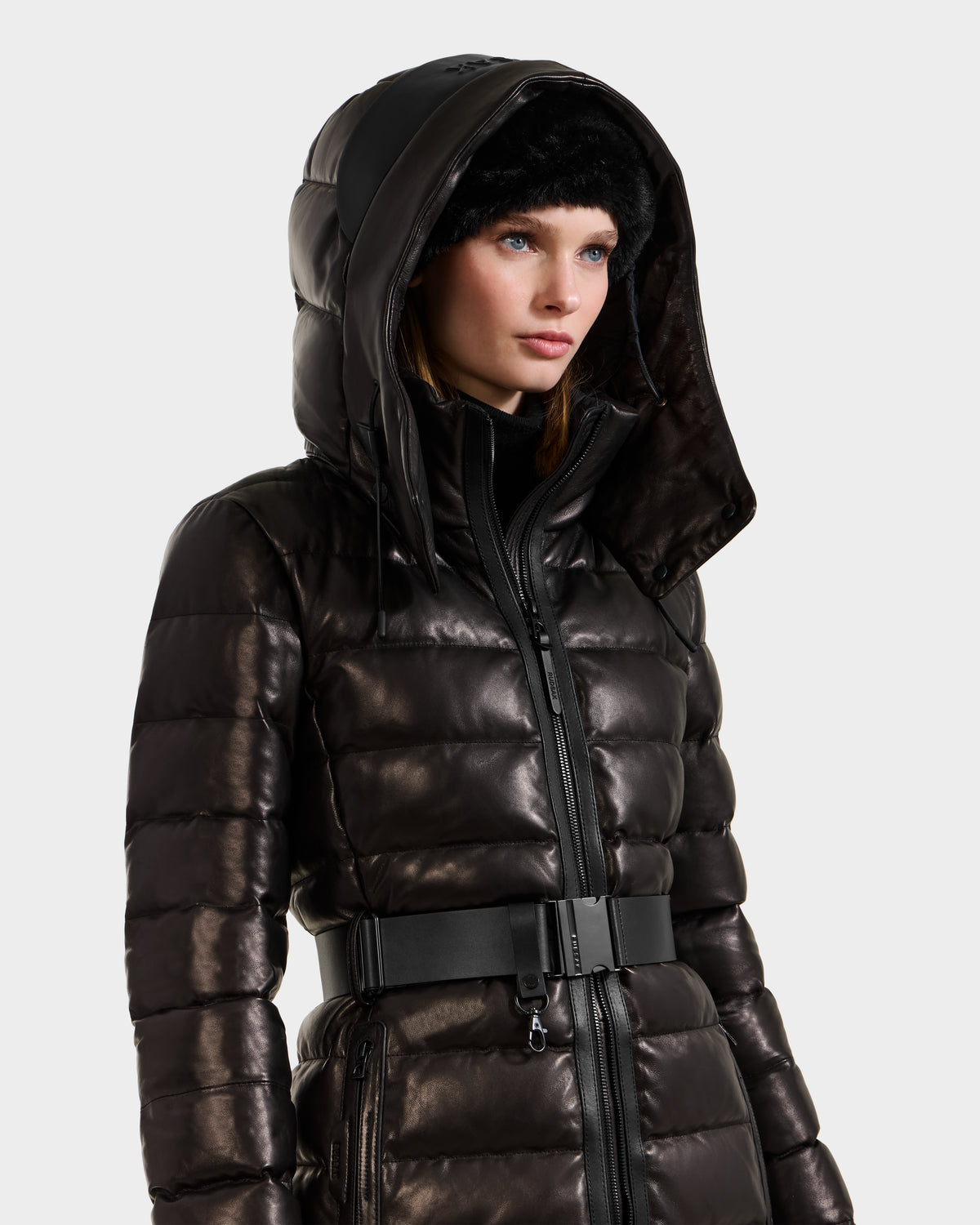 A woman with straight hair and blue eyes wears the Rudsak PELIN X - 8125575 Black leather down puffer with a hood and black belt, paired with a black headband, standing against a light plain background.