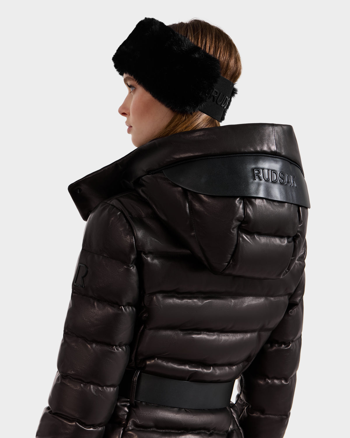 A woman in the PELIN X - 8125575 Black down puffer jacket by Rudsak, featuring a shiny black leather finish, hood, and wide collar, wears a black Rudsak fur headband. She looks to the side against a plain white background.