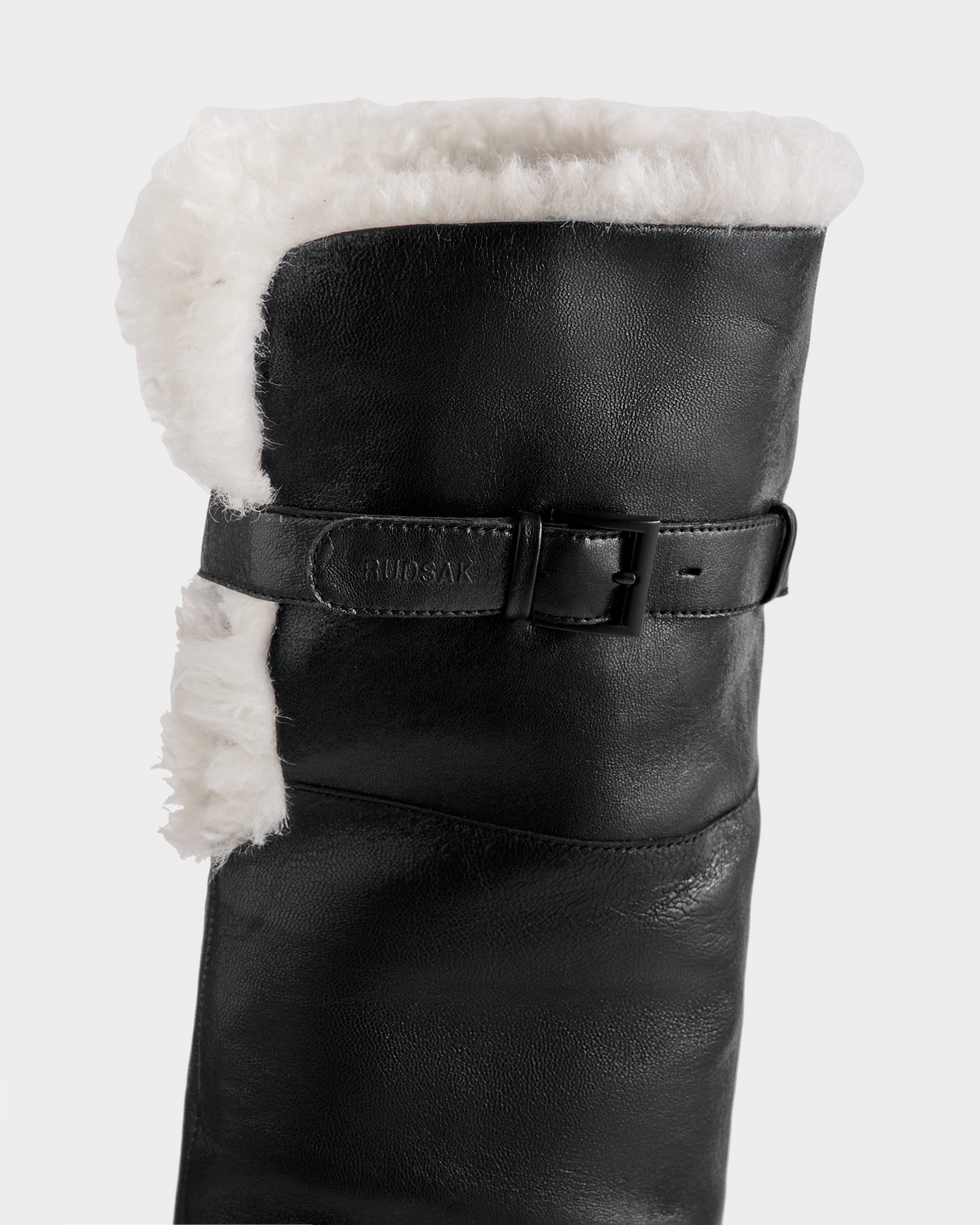 A close-up of Rudsak's PERNA - 8225555 Black women's winter boots, showcasing black leather, a white shearling lining, and a decorative side strap with buckle.